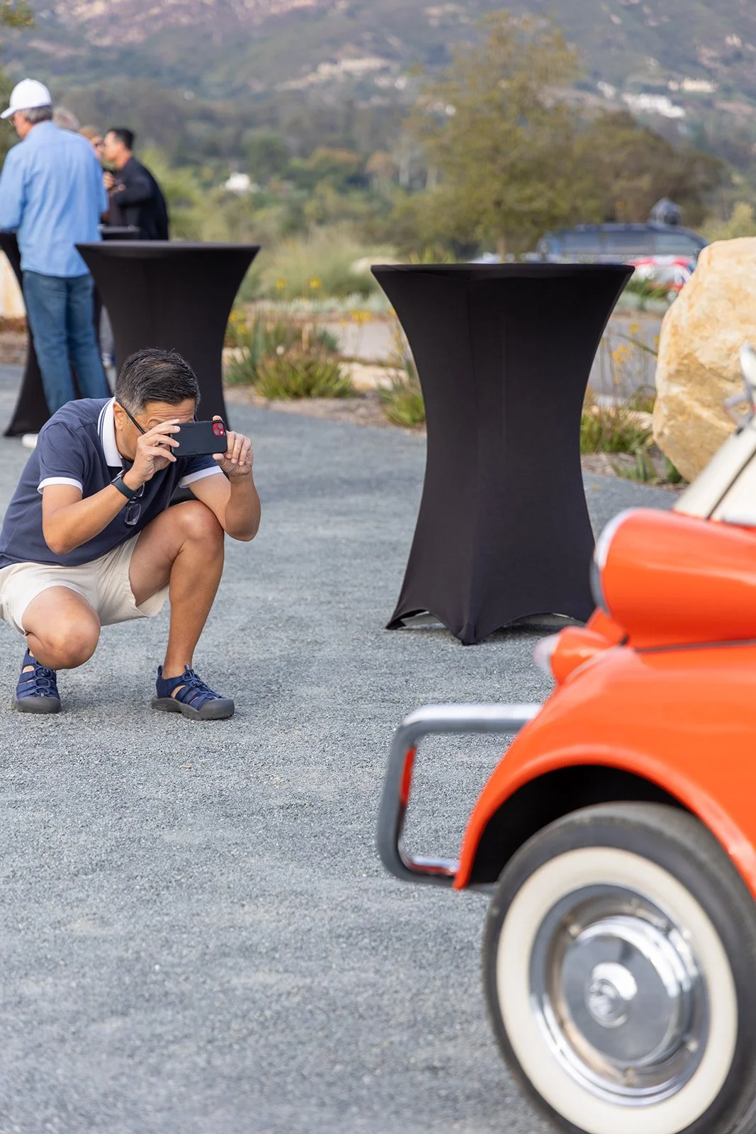 A man squatting on the ground taking a photo with his phone at an outdoor event, with a classic orange car and several high-top tables covered in black cloths in the background.