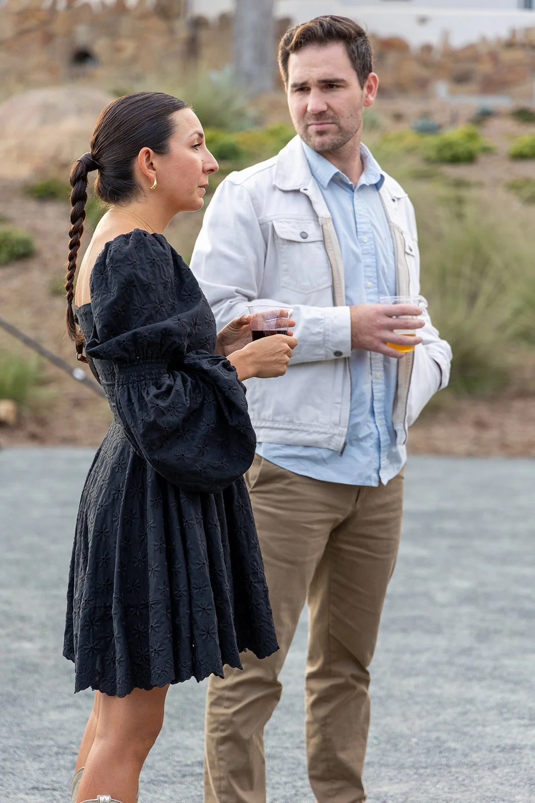 A woman in a black dress with puffed sleeves holds a glass of red wine, and a man in a light blue shirt and beige pants holds a glass of beer, standing outdoors on a gravel surface with a scenic background.
