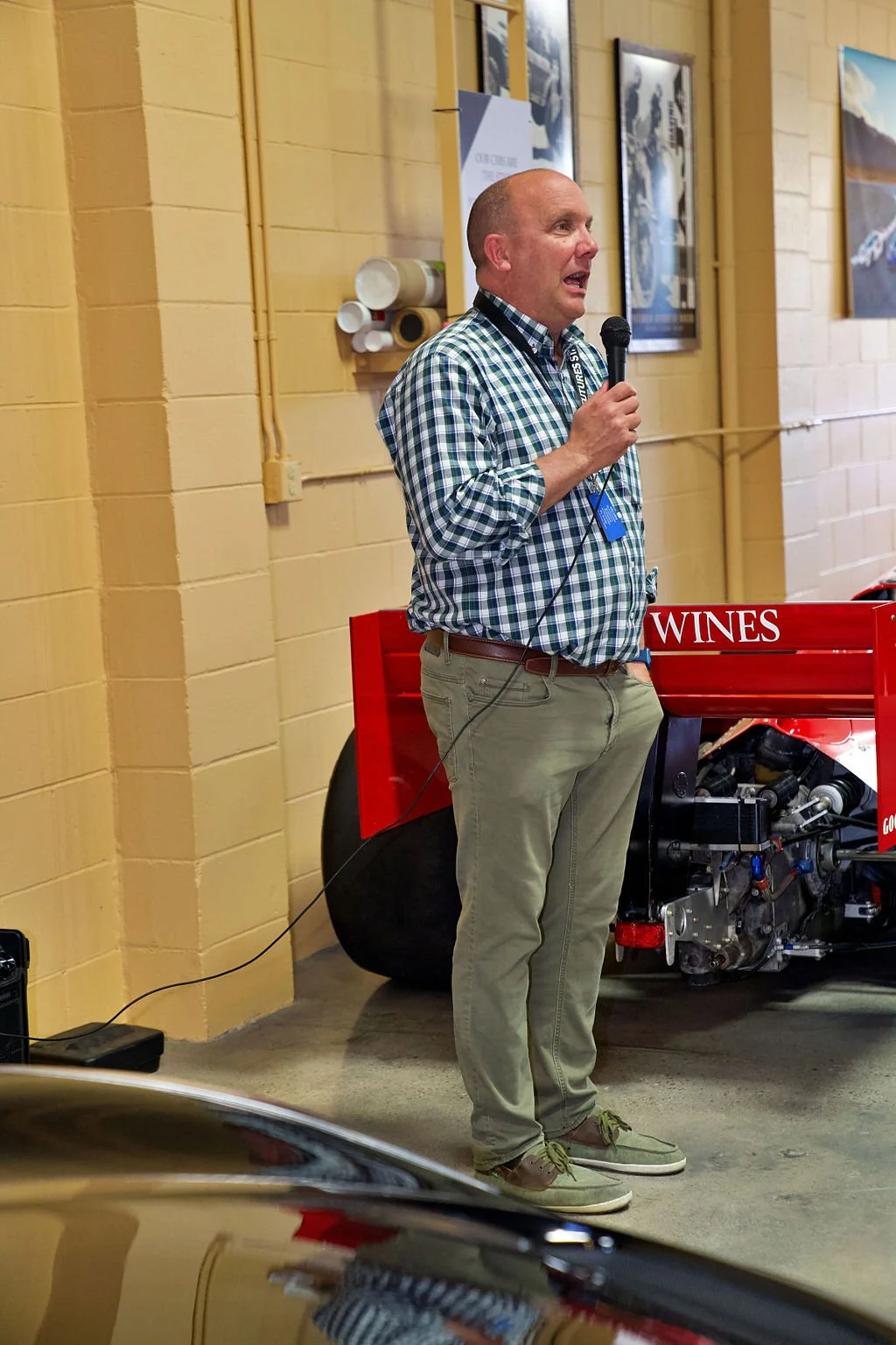 A man with a plaid shirt holding a microphone, speaking in front of a vintage race car with a red and black color scheme, inside a garage or showroom with framed posters on the wall.