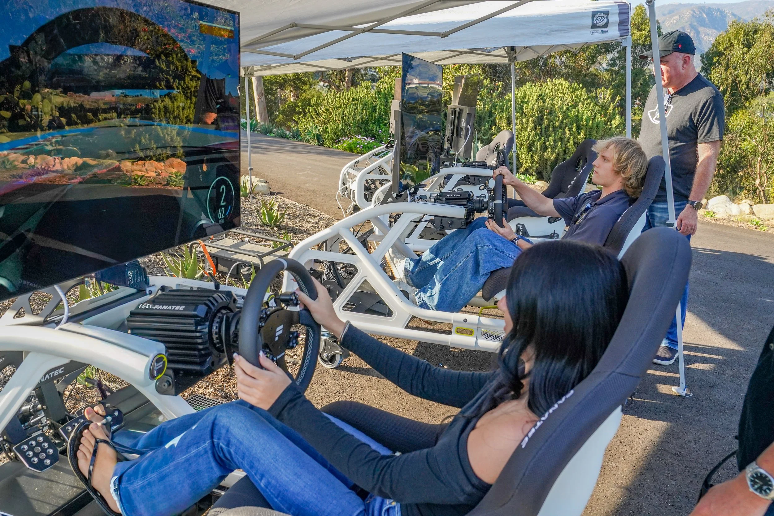 People participating in a racing simulation experience outdoors with racing seats, steering wheels, and multiple monitors, under a white canopy on a sunny day with green shrubbery and mountains in the background.