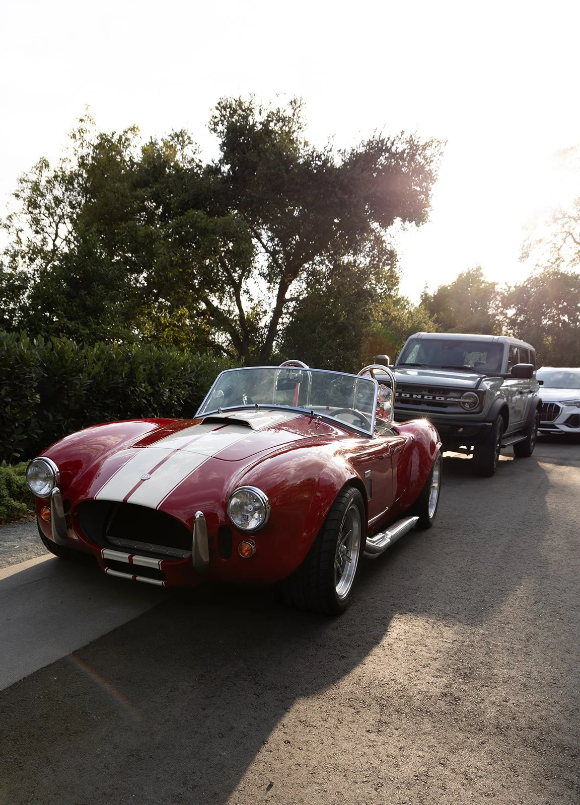 A red vintage sports car with white racing stripes parked on the side of the road, with a modern gray SUV and a white sports car behind it, in a natural setting with trees and sunlight.