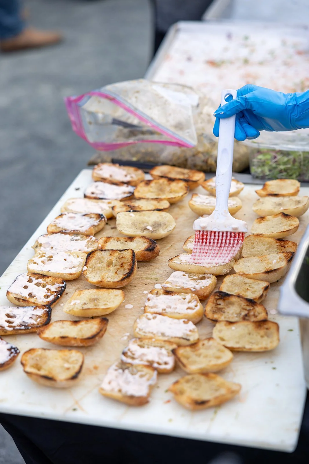 Person in blue gloves spreading a creamy topping on grilled slices of baguette at a street food stand.