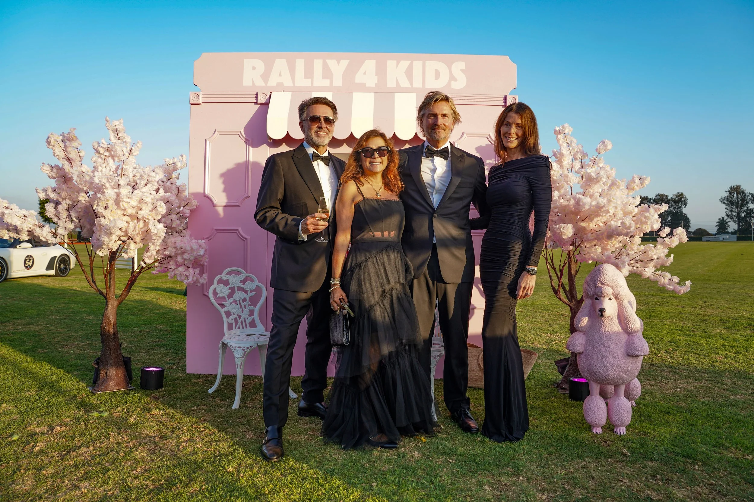 Group of five people in formal attire standing in front of a pink backdrop with a sign that reads 'Rally 4 Kids,' decorated with pink cherry blossom trees and a pink poodle statue on a grassy field.
