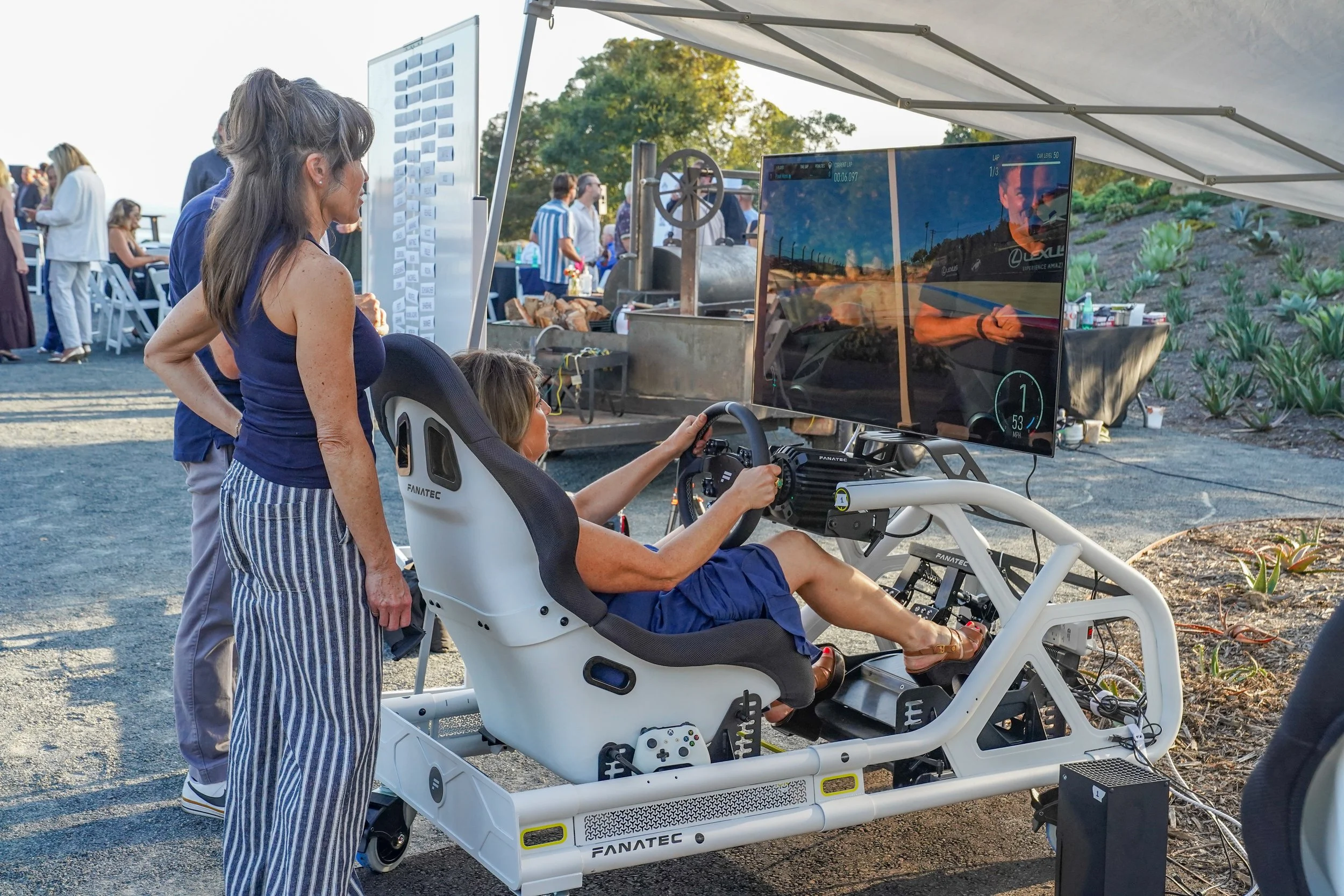 A woman sits in a racing simulator with a large screen displaying a racing game, while another woman stands beside her, observing. There are people in the background at an outdoor event with trees and tables.
