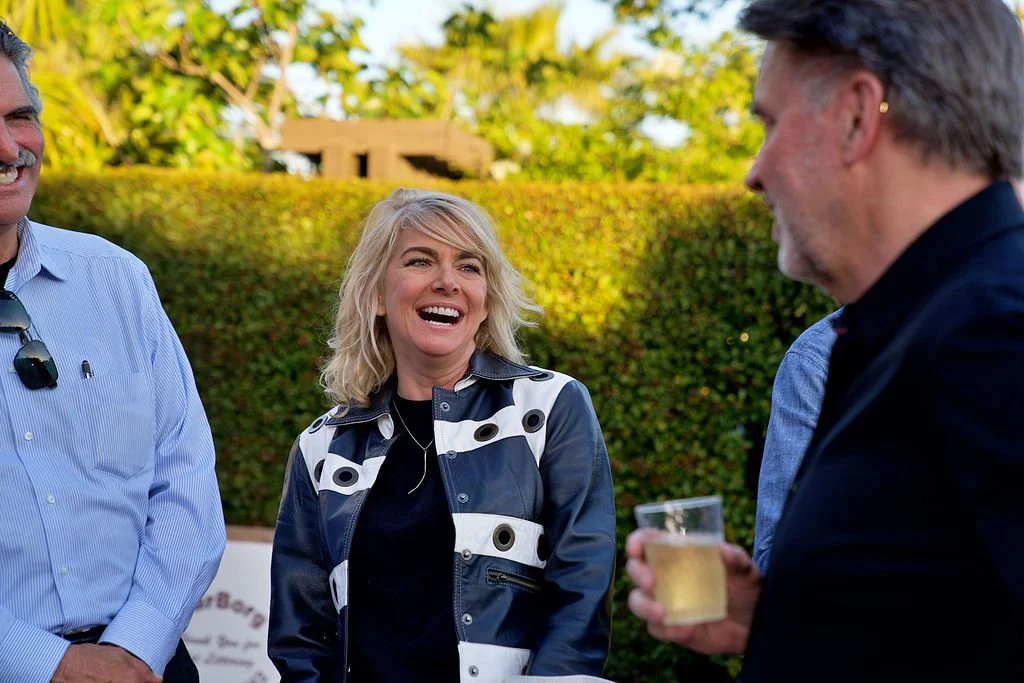 People smiling and talking outdoors at a social gathering, with a woman in a patterned jacket holding a drink.