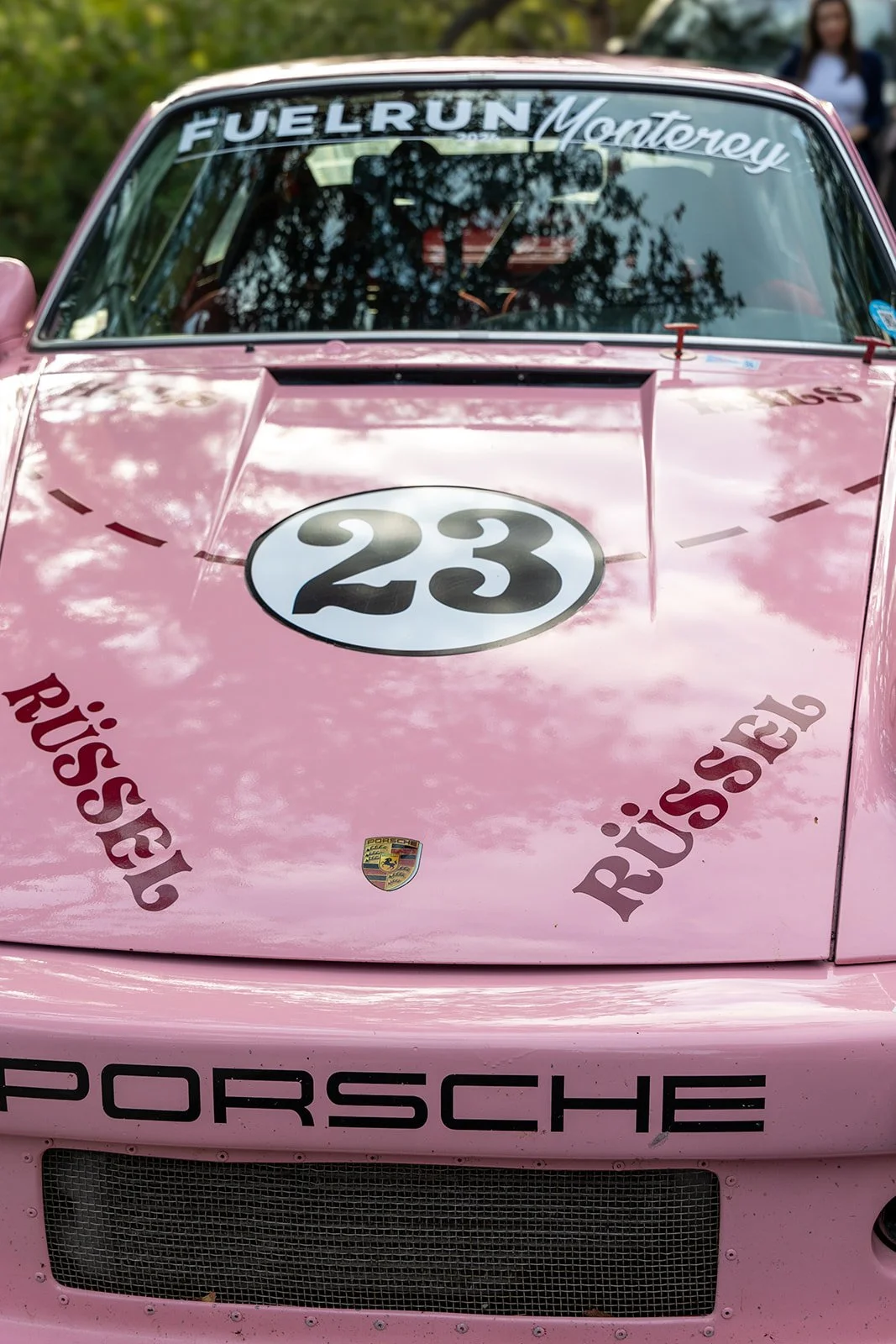 A pink Porsche race car with the number 23 on the hood, featuring decals for 'Rüssel', 'Fuel Run Monterey', and a Porsche emblem, parked outdoors with trees reflected on the windshield.