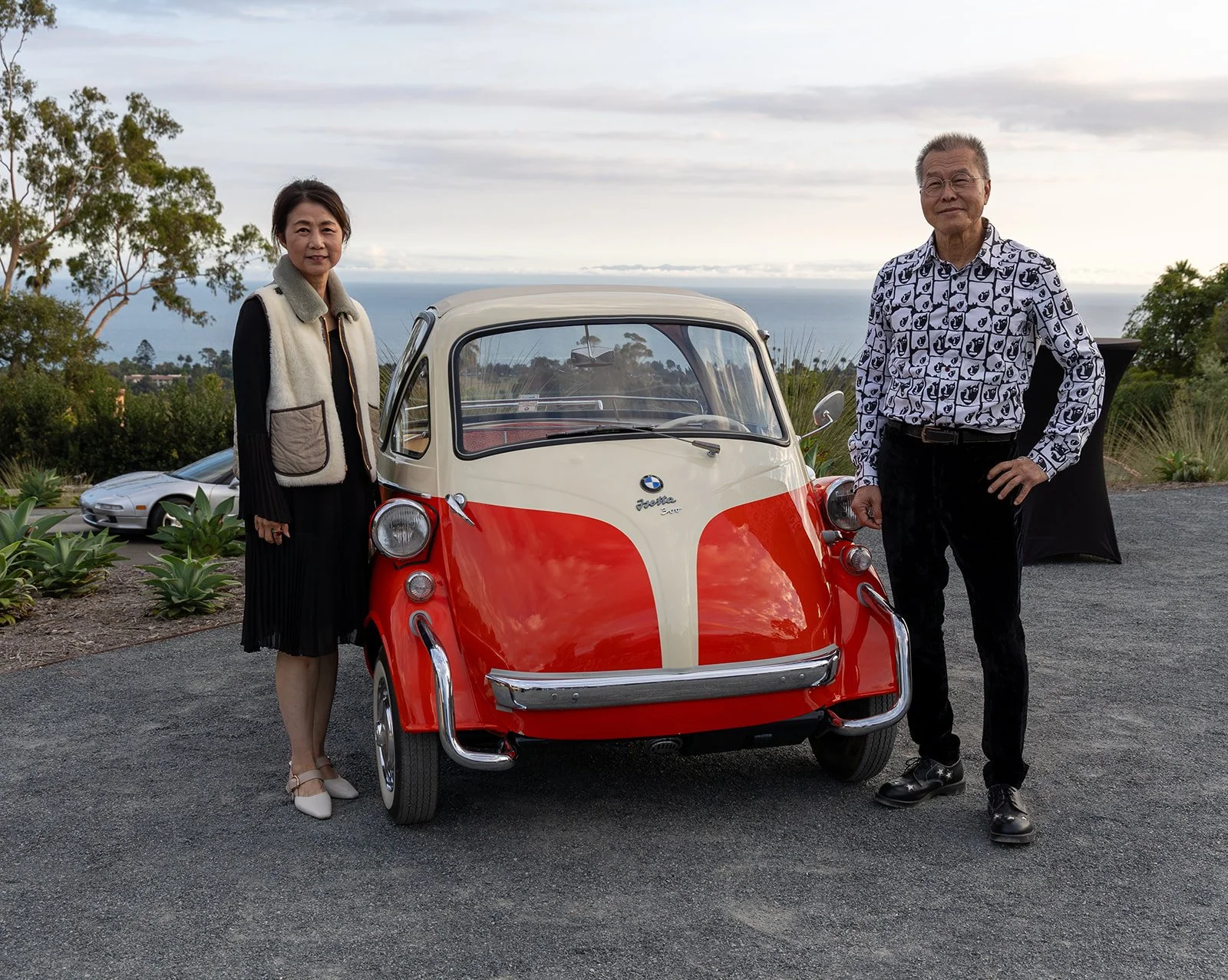 A woman and an older man stand next to a red and cream BMW Isetta microcar outdoors with the ocean and cloudy sky in the background. The woman is wearing a black dress with a beige vest, and the man is wearing a patterned shirt and black pants.