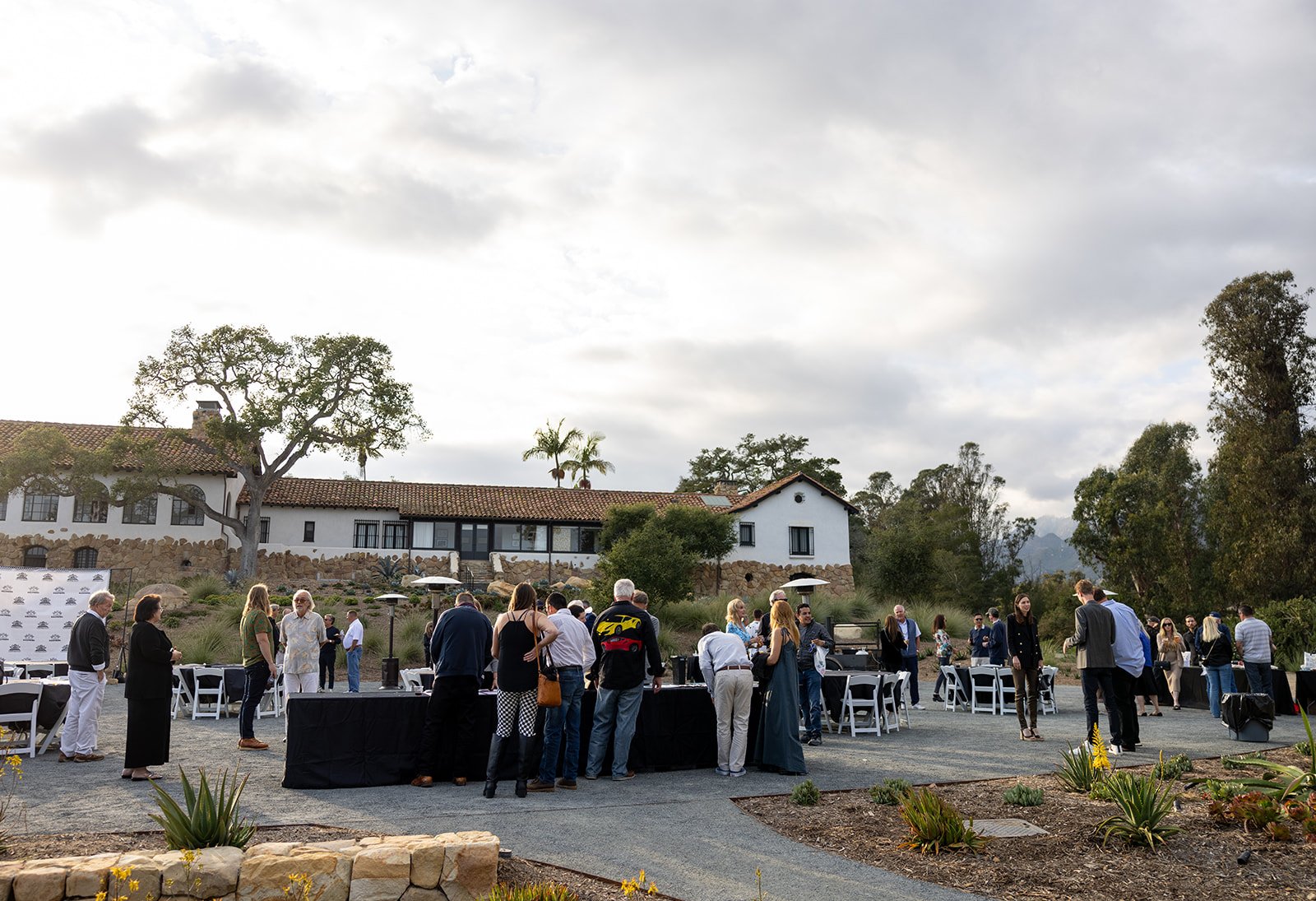 People socializing outdoors at a courtyard event with tables, chairs, and umbrellas, with a large white house, trees, and a cloudy sky in the background.