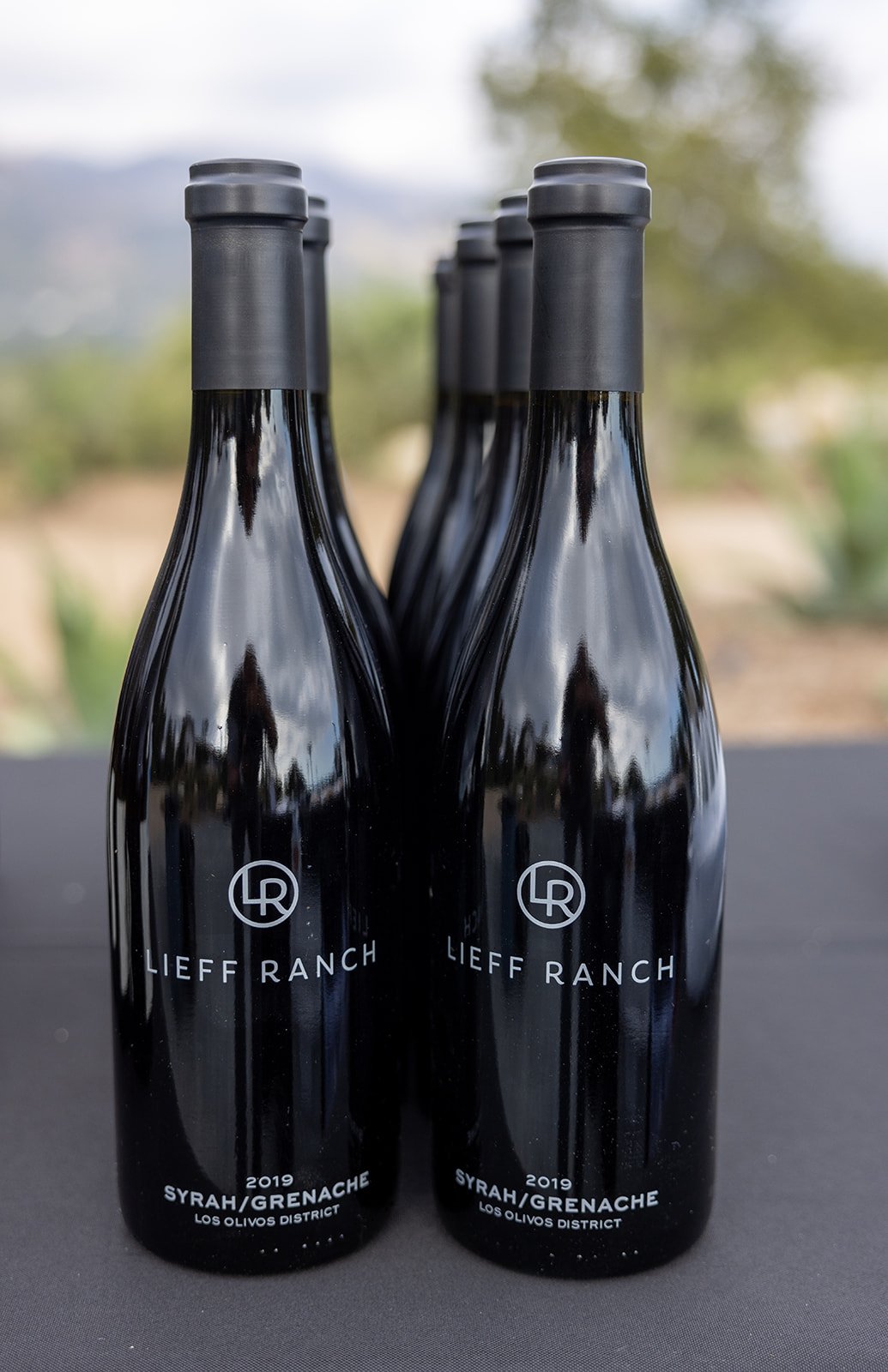 Four black wine bottles with gray caps, labeled "Lieff Ranch 2019 Syrah/Grenache, Los Olivos District," arranged in two rows against a blurred outdoor background.