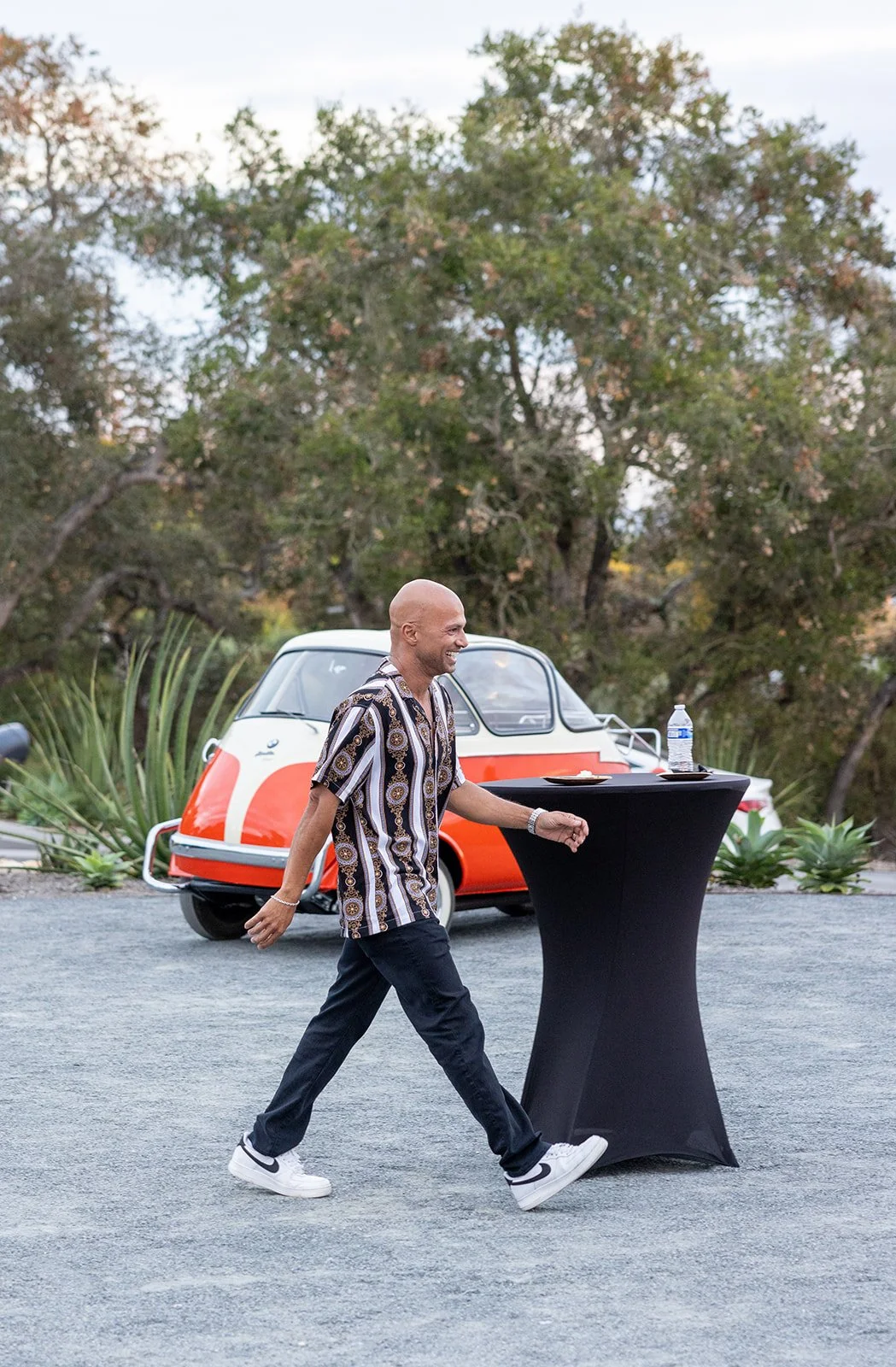 A smiling man in a patterned short-sleeve shirt, black pants, and white sneakers walking past a tall black cocktail table with a water bottle and plate on it, outdoors near a red and white vintage microcar and green trees.