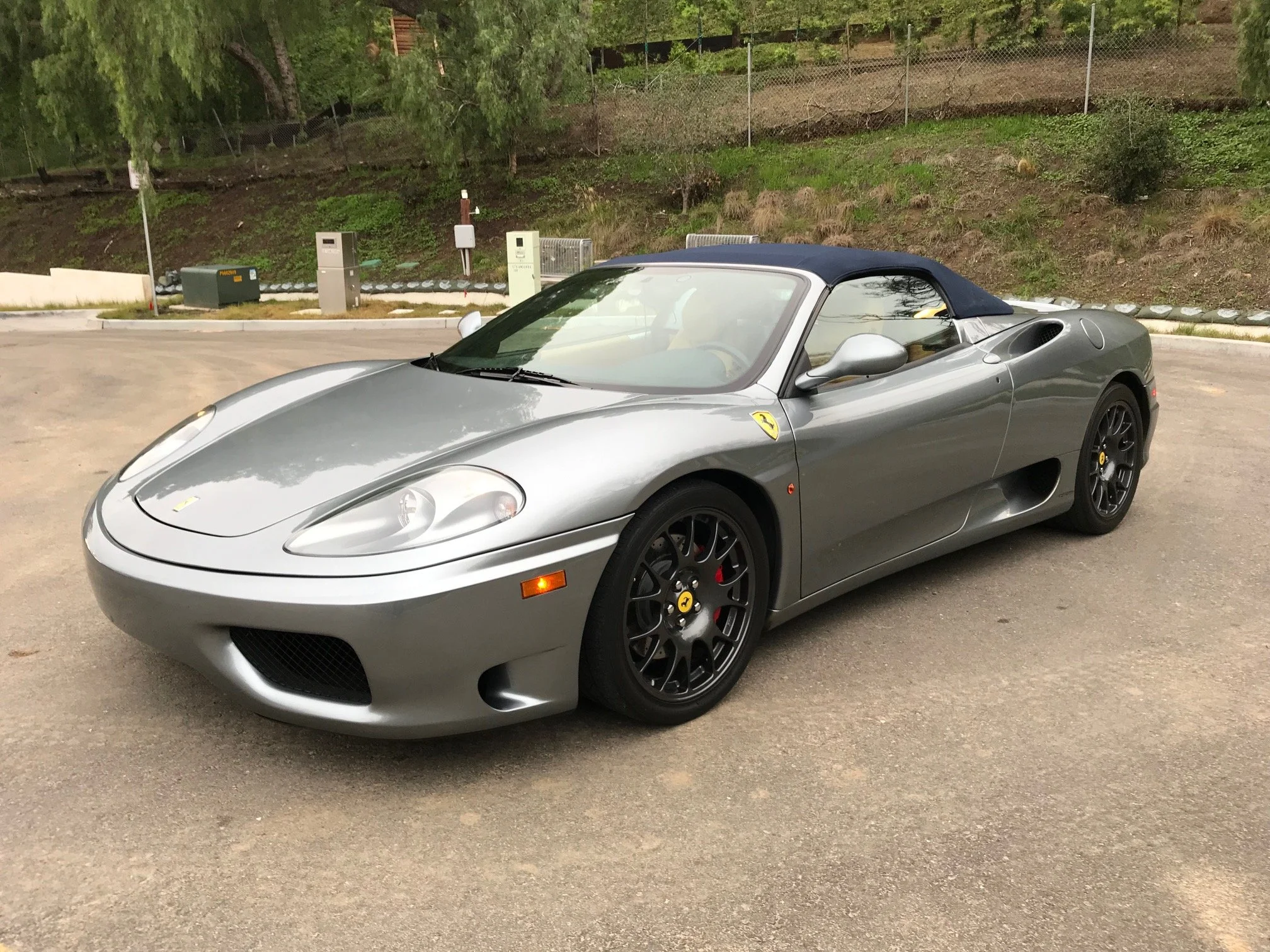 Silver Ferrari sports car with black wheels parked on a paved area with trees and a fence in the background.