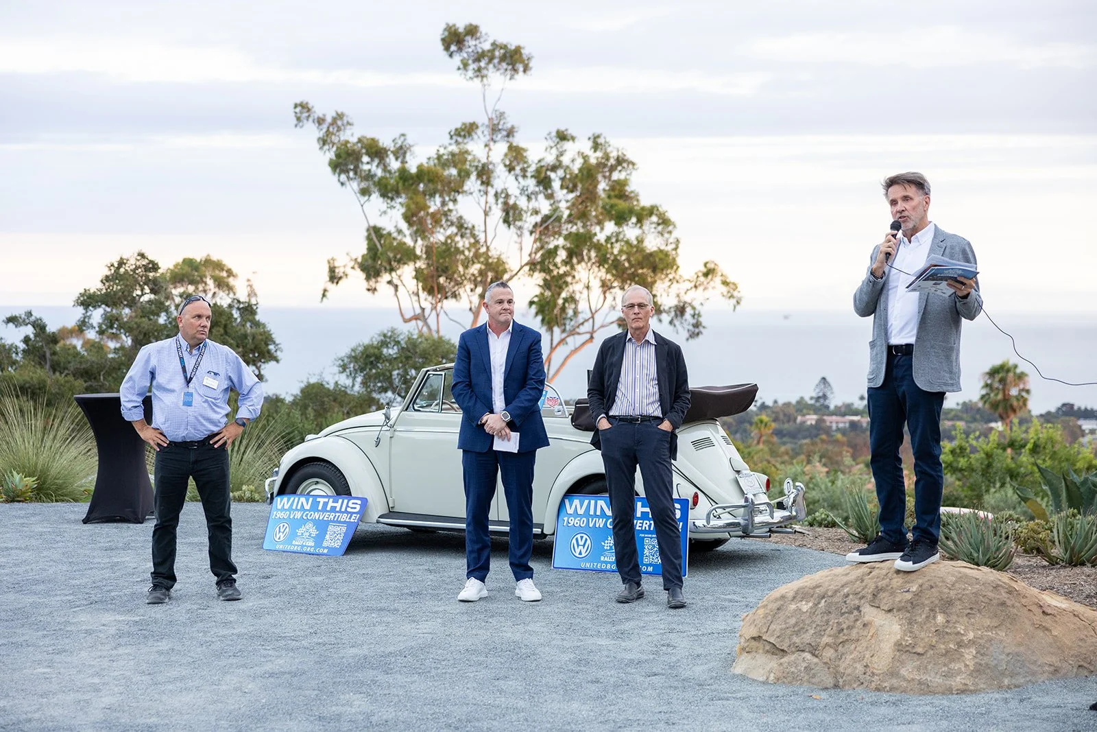 Four men standing outdoors near a vintage white Volkswagen convertible car, with donation signs, trees, and a scenic view in the background, one man speaking into a microphone on a rock.