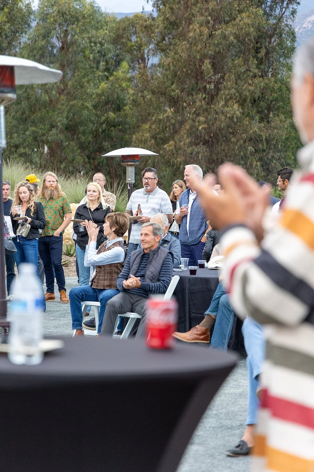 People gathered outdoors at a social event, some seated and others standing, with trees and mountains in the background. Various individuals are holding drinks and engaging in conversations.