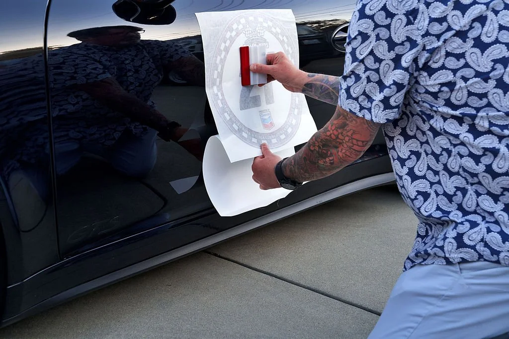 Person with tattoos on their right arm applying a decal to a black car's side door using a squeegee.