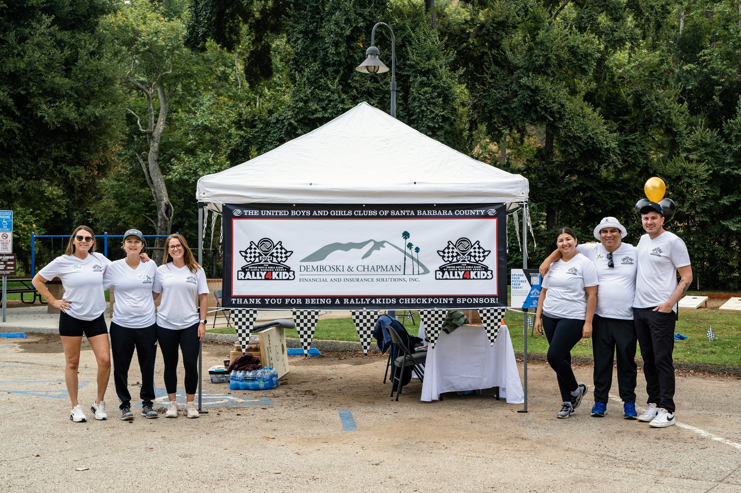 Group of six people standing in front of a tent at an outdoor event, with a sign reading "Rally 4 Kids" and sponsorship information from Demboski & Chapman Financial and Insurance Solutions, Inc., organized by the United Boys and Girls Clubs of Santa