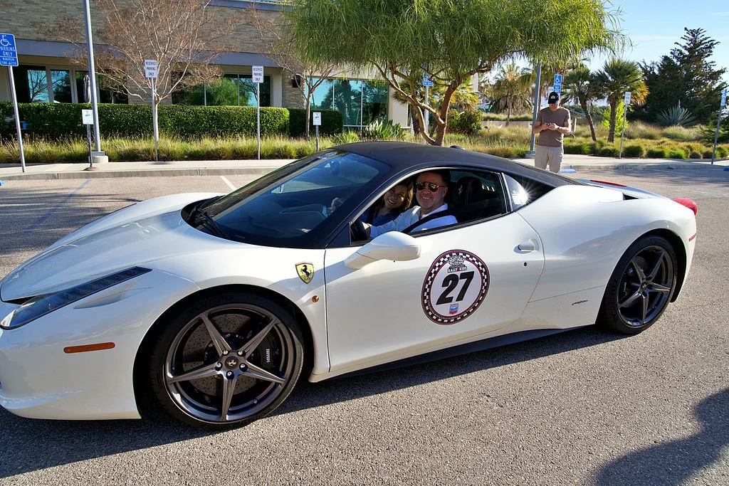 A white sports car with a Ferrari badge and a racing number 27 decal on the door, parked in a lot. Two people are seated inside, smiling. A man is standing outside nearby, looking at his phone, with trees and a building in the background.