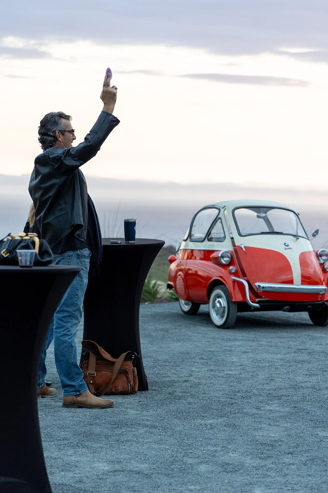 A man in a leather jacket holding up a peace sign with his right hand, standing outdoors near a small red and white microcar, with a table with a drink and a bag on the ground nearby. The sky is cloudy, and the scene appears to be at dusk or dawn.