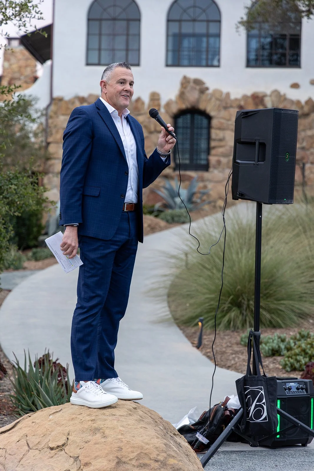A man in a blue suit holding a microphone and a piece of paper, speaking outdoors next to a speaker, with a building and plants in the background.