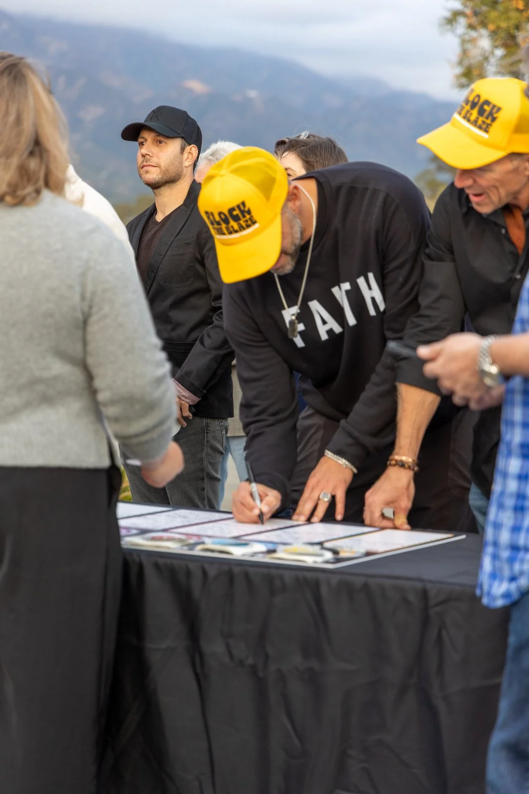 Group of people gathered outdoors, signing posters or photos on a table, with mountains in the background. Some individuals wear yellow caps with black text that reads 'BLOCK THE BLAZE'.