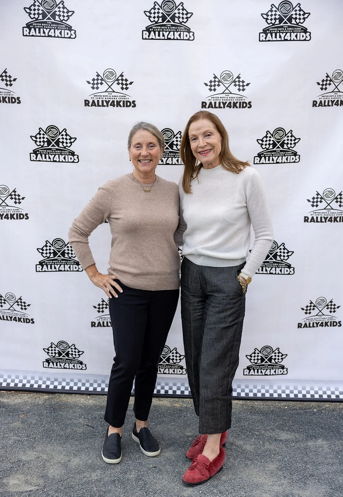 Two women standing in front of a Rally4Kids event backdrop featuring a checkered flag and racing emblem, smiling at the camera.