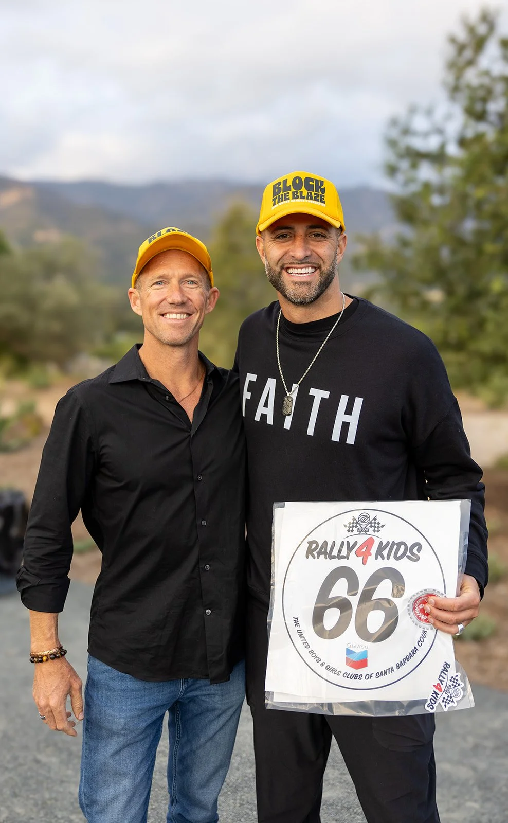Two smiling men standing outdoors. The man on the right is holding a sign with the number 66 and the text 'Rally4Kids,' wearing a yellow cap with 'BLOCK THE BLAZE' written on it, a black shirt, and a chain necklace. The man on the left is wearing a y