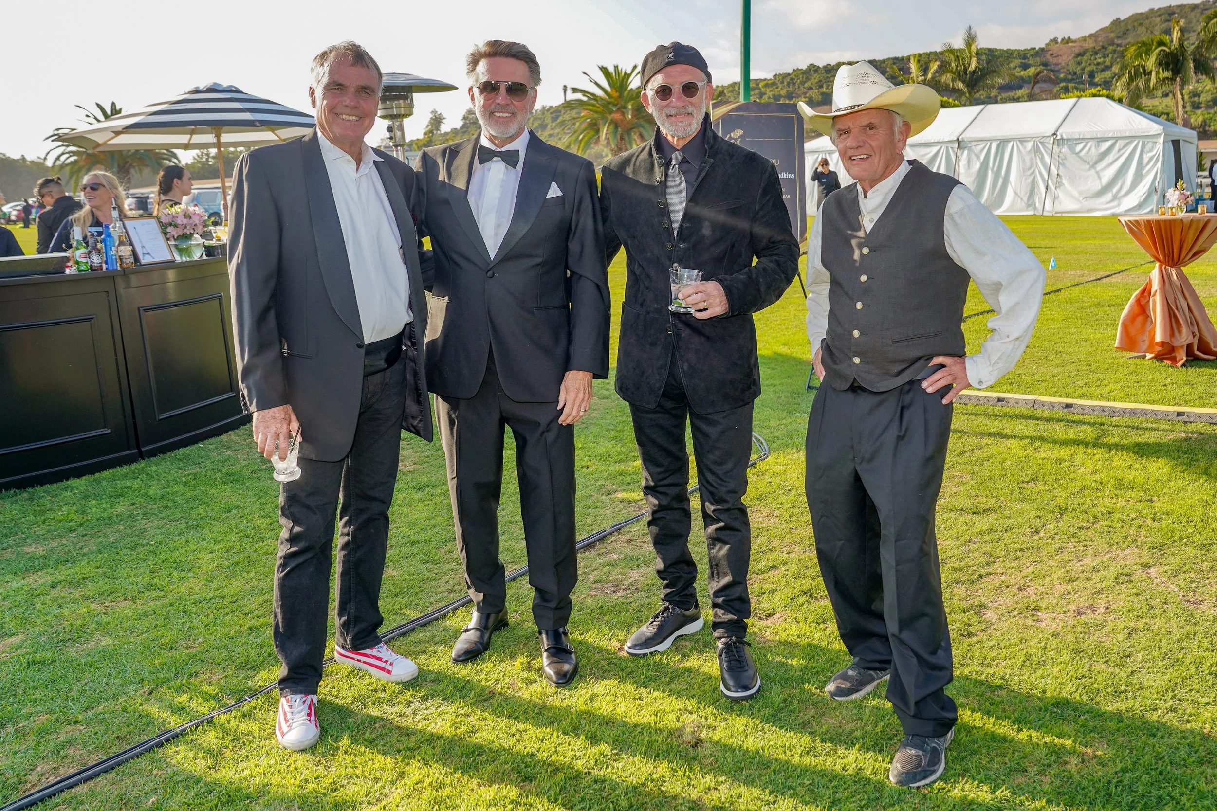 Four men dressed in formal and casual attire standing on grass at an outdoor event, with palm trees and event tents in the background.