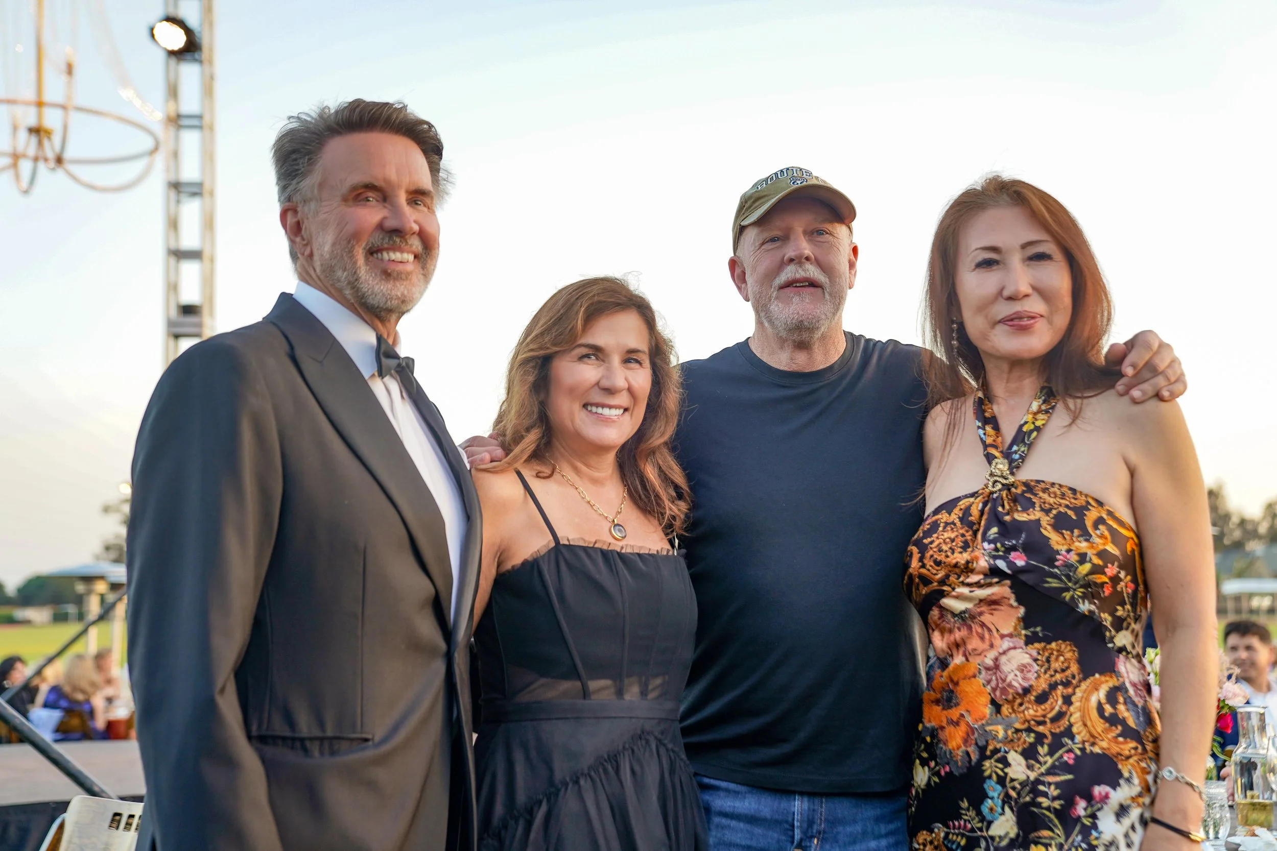 Group of four people smiling and posing outdoors at an event during daytime. One man is in a tuxedo, another man is wearing a baseball cap and casual clothes, a woman is in a black dress, and another woman is in a floral dress. They are standing clos