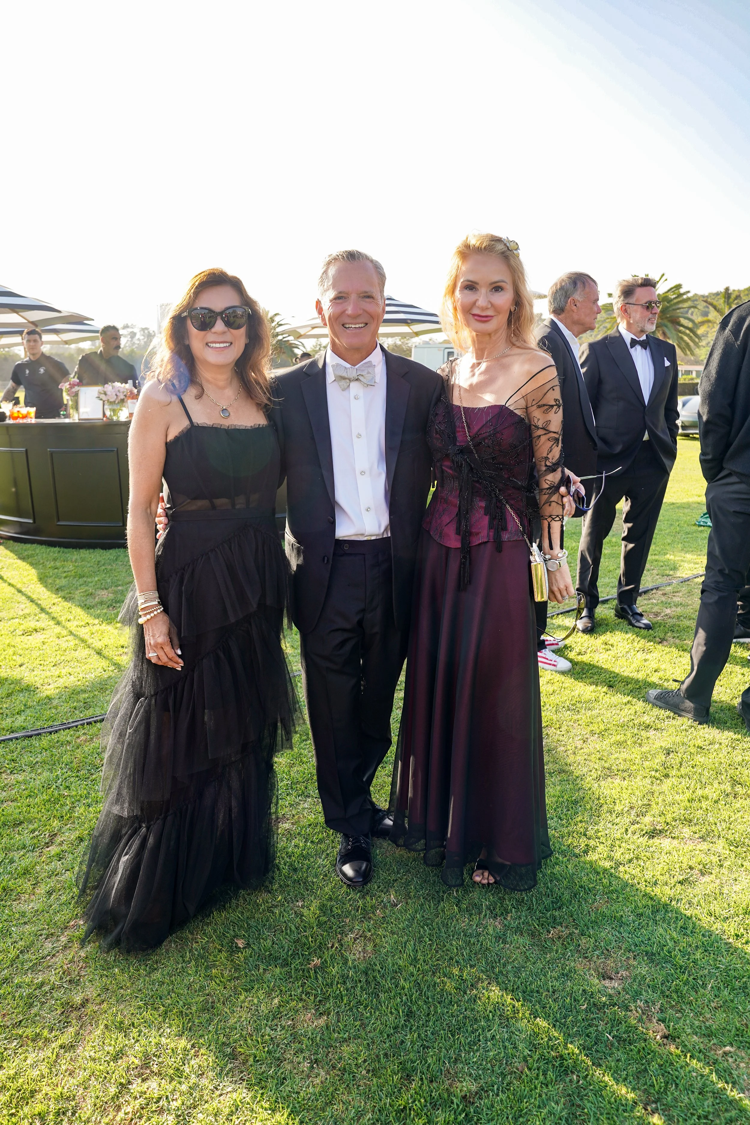 Three people dressed in formal attire standing on grass at an outdoor event, with a bar and other guests in the background.