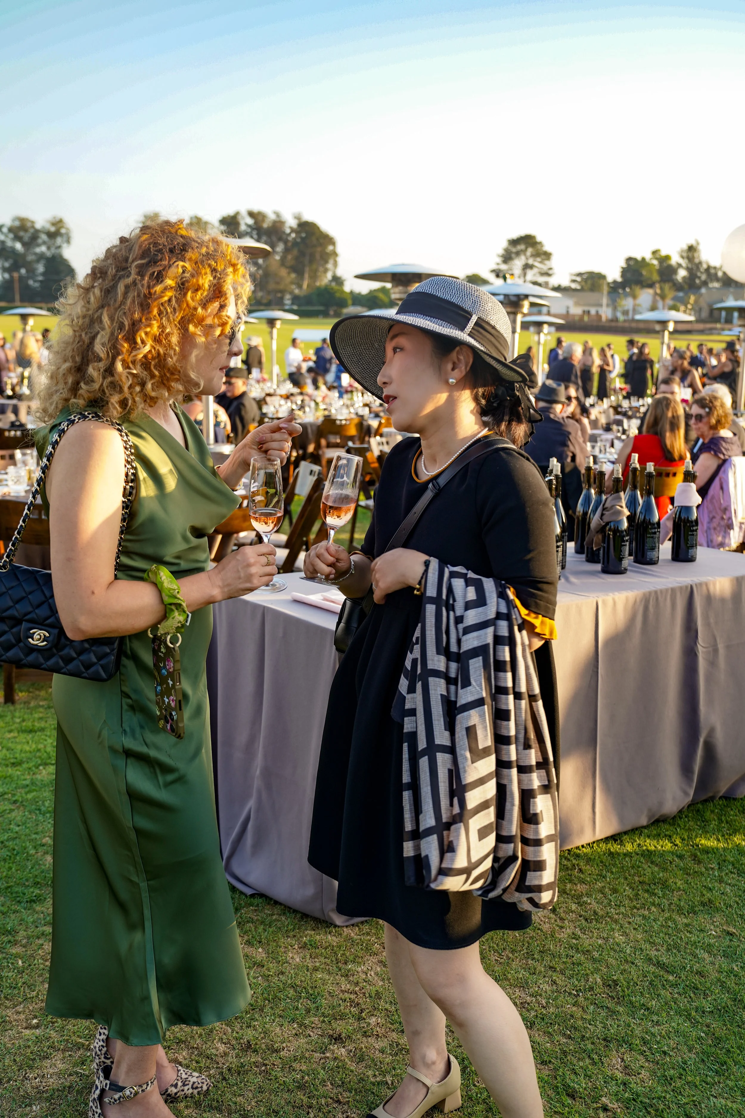 Two women talking at an outdoor event, each holding a glass of rosé wine. One woman has curly red hair and is wearing a green dress with a Chanel purse, while the other has dark hair with a wide-brimmed hat, black dress, and patterned bag. There are 