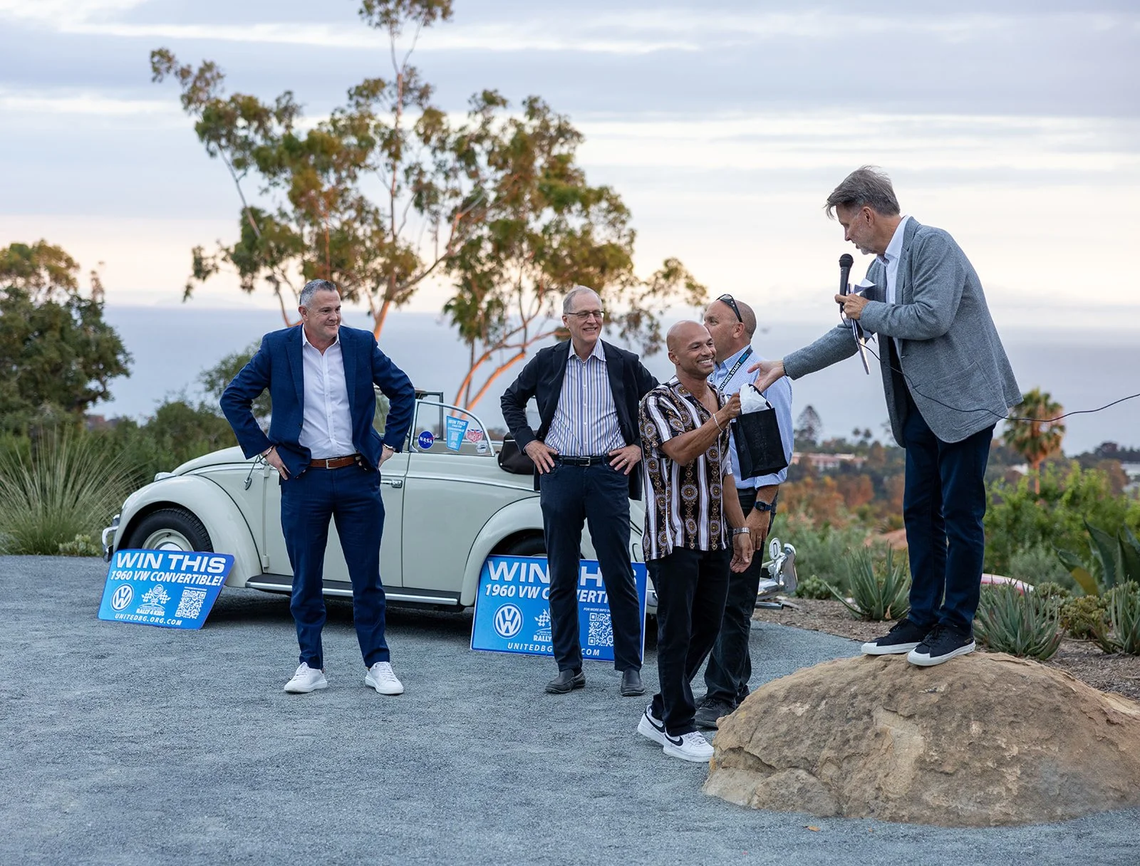 A group of five men standing outdoors near a vintage white Volkswagen convertible car, with signs promoting a contest to win the car, on a scenic hilltop with trees and ocean in the background. One man is standing on a rock, speaking into a microphon