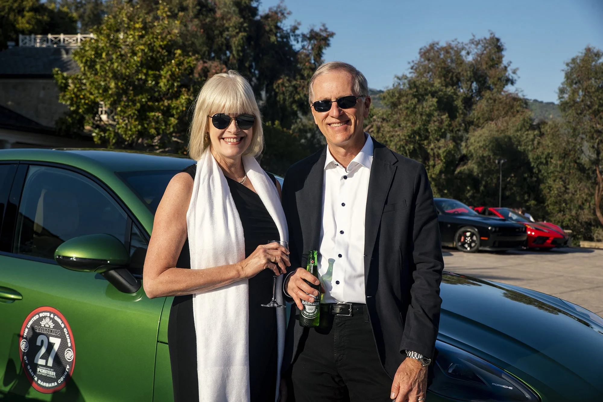 A smiling couple wearing sunglasses at a car event, standing in front of a green Porsche with a rally badge, holding drinks, with other sports cars in the background under a clear blue sky.