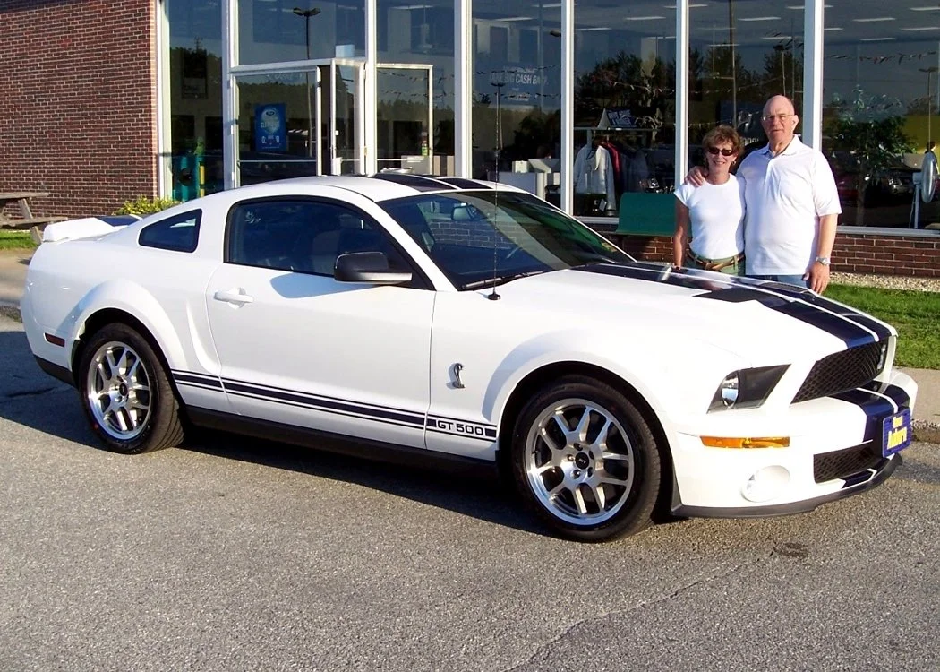A white Ford Mustang Shelby GT500 with black racing stripes parked outside a car dealership, with a smiling older couple standing behind it.