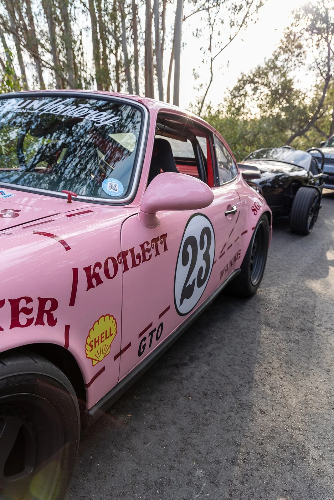 Pink race car with the number 23 and various sponsor logos, parked outdoors among trees.