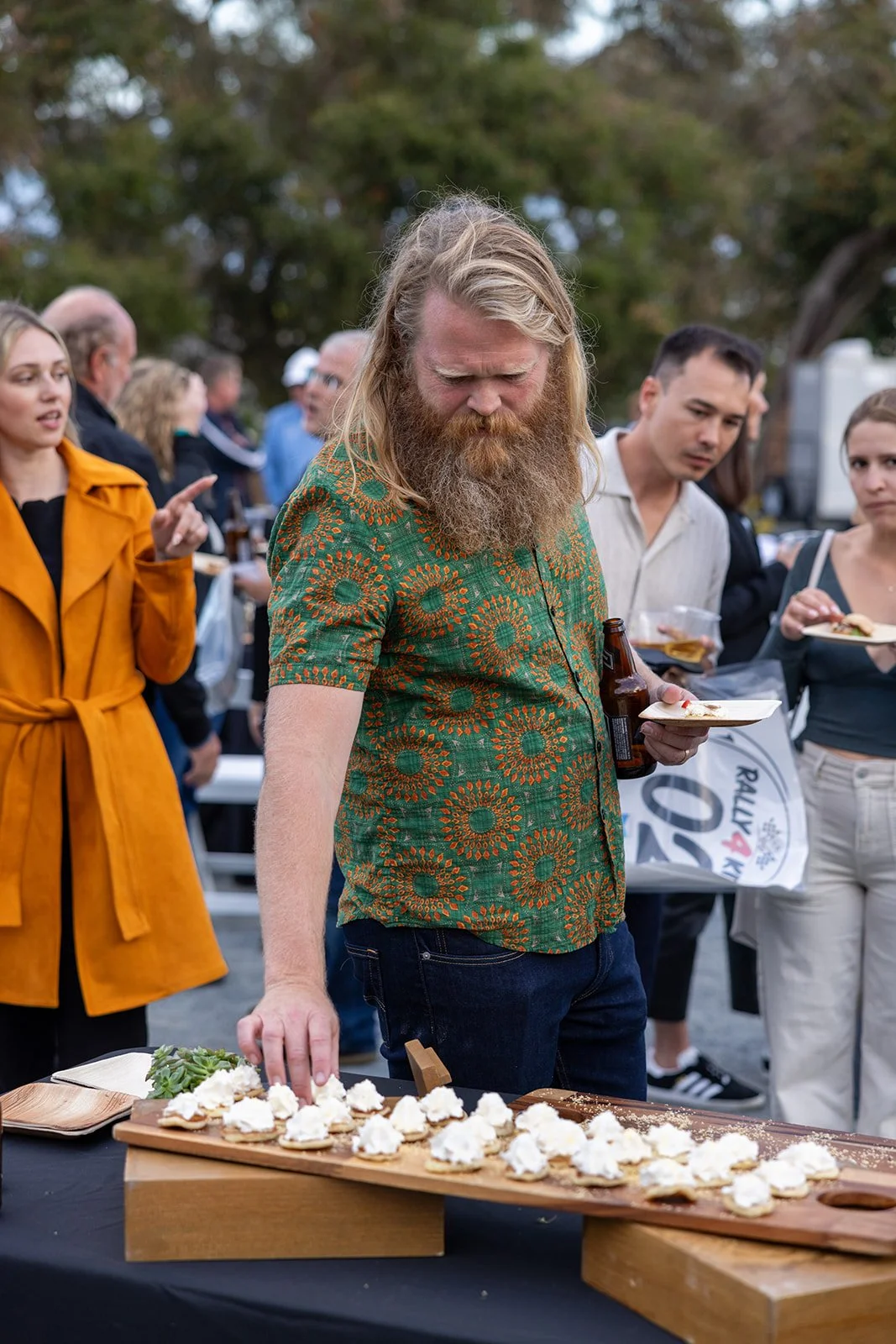 A man with long blonde hair and a beard wearing a green patterned shirt is reaching for food on a wooden platter, at an outdoor gathering with several people around him, some holding plates and drinks.