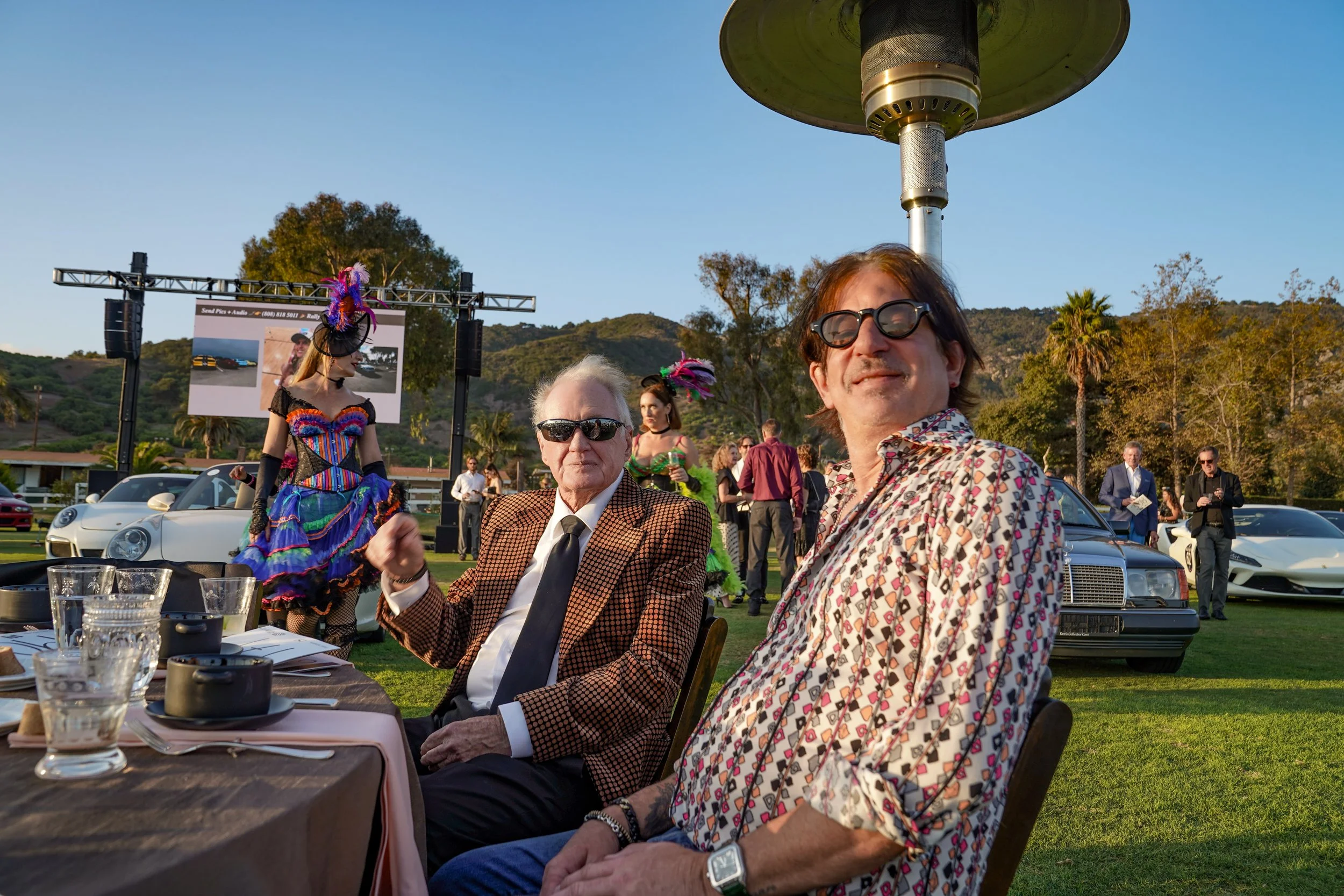 Two men sitting at a table outdoors at a car event, with luxury cars and people in the background, and a person in a colorful outfit and hat dancing nearby.