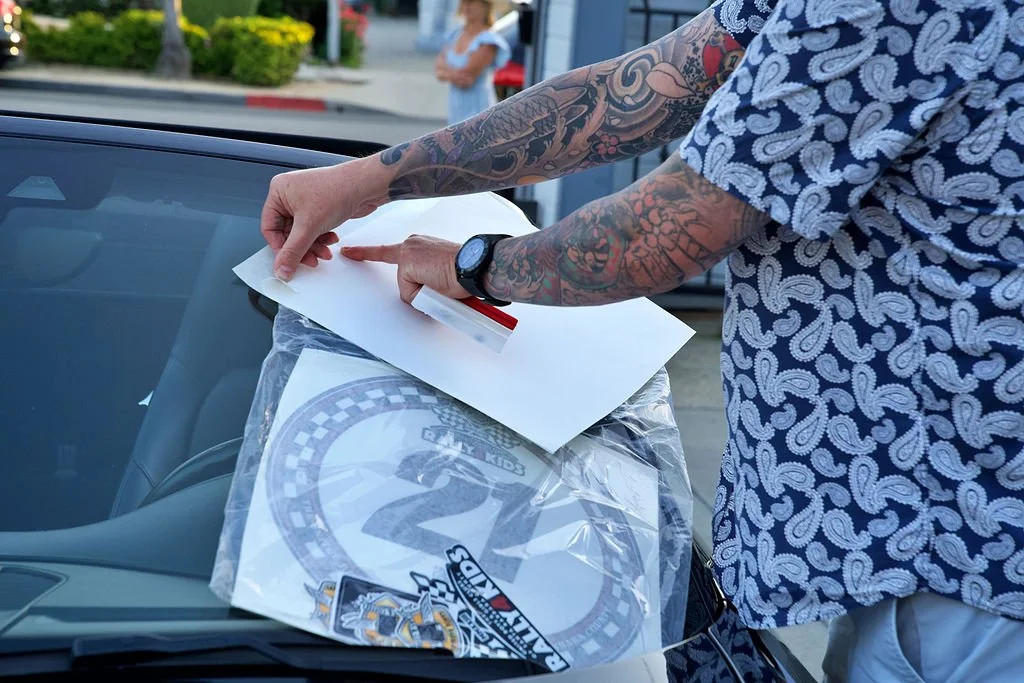 A person with arm tattoos is sticking a white paper into a plastic bag containing a children's race bib with the number 2 on it, placed on the hood of a parked car.