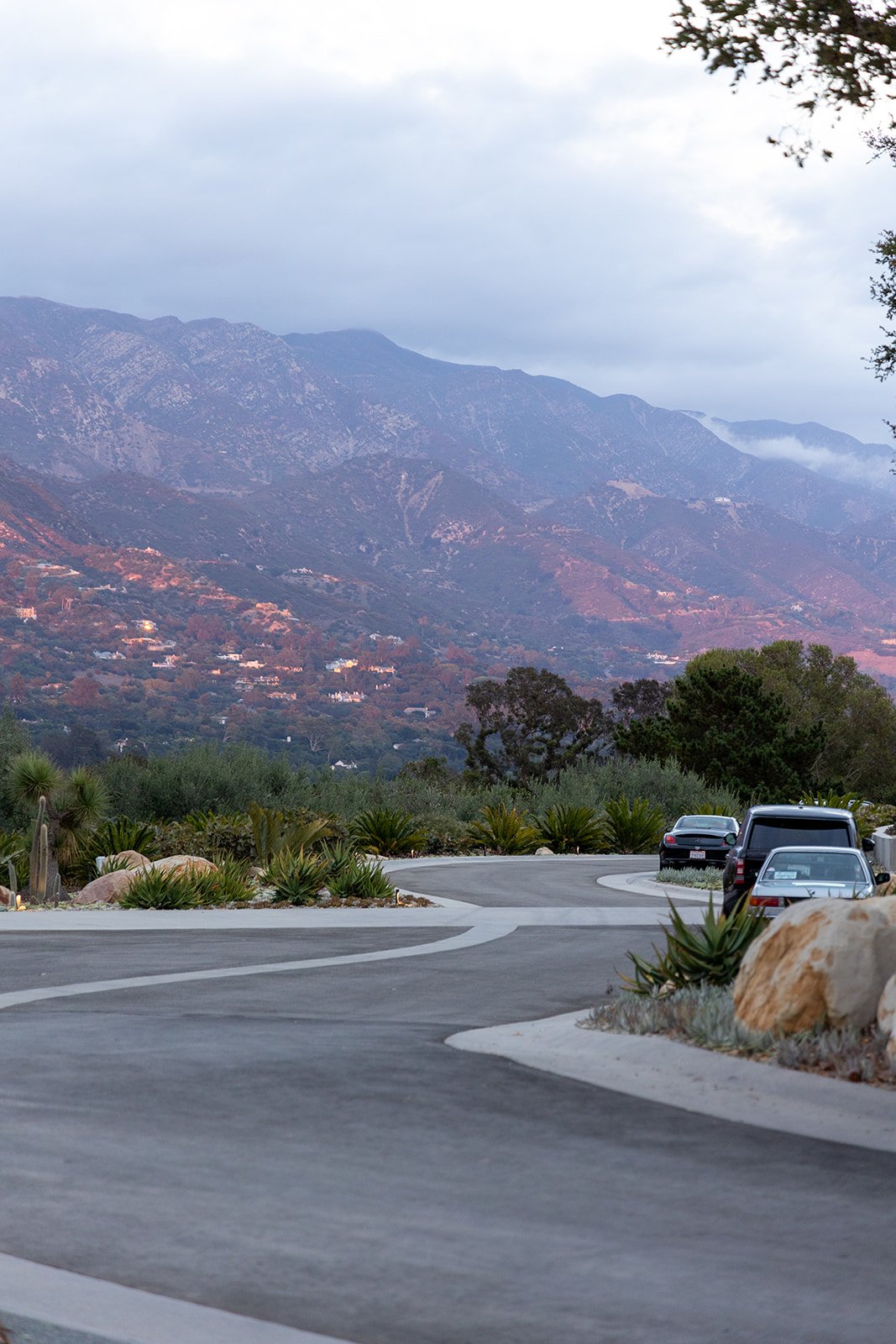 A winding paved road with cars parked along the side, surrounded by rocks and desert plants, with a mountain range in the background under a cloudy sky.