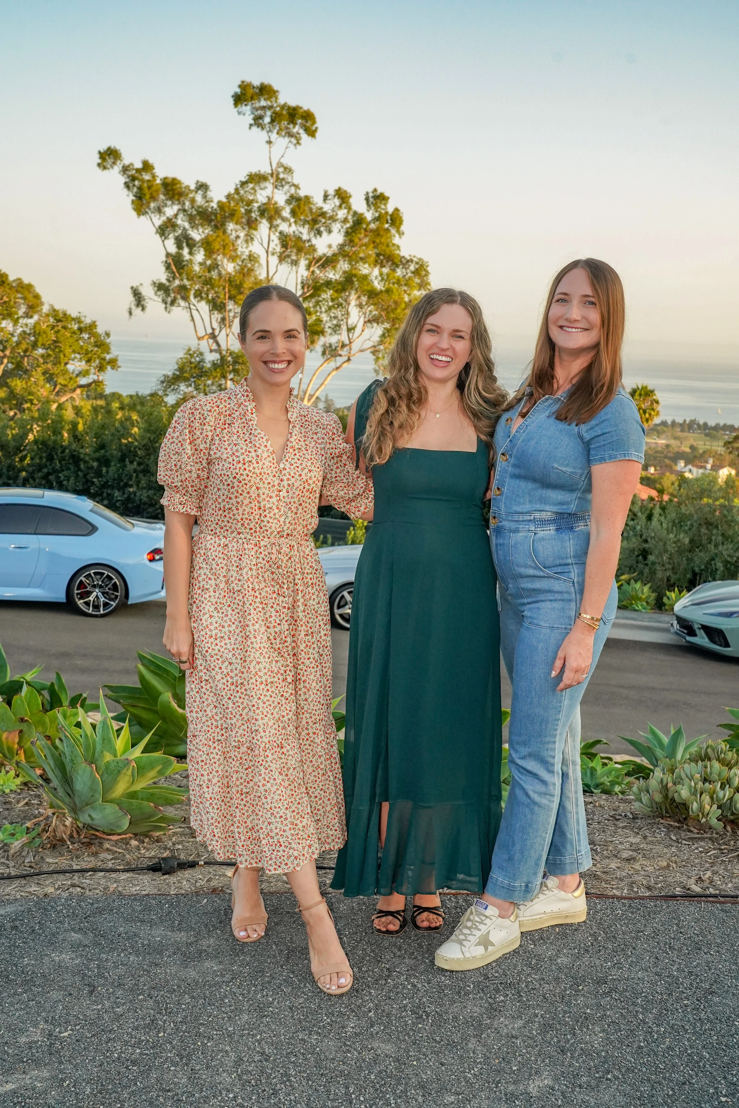 Three women standing outdoors on a hill with a view of the ocean, trees, and parked cars in the background, smiling and dressed in casual summer clothing.