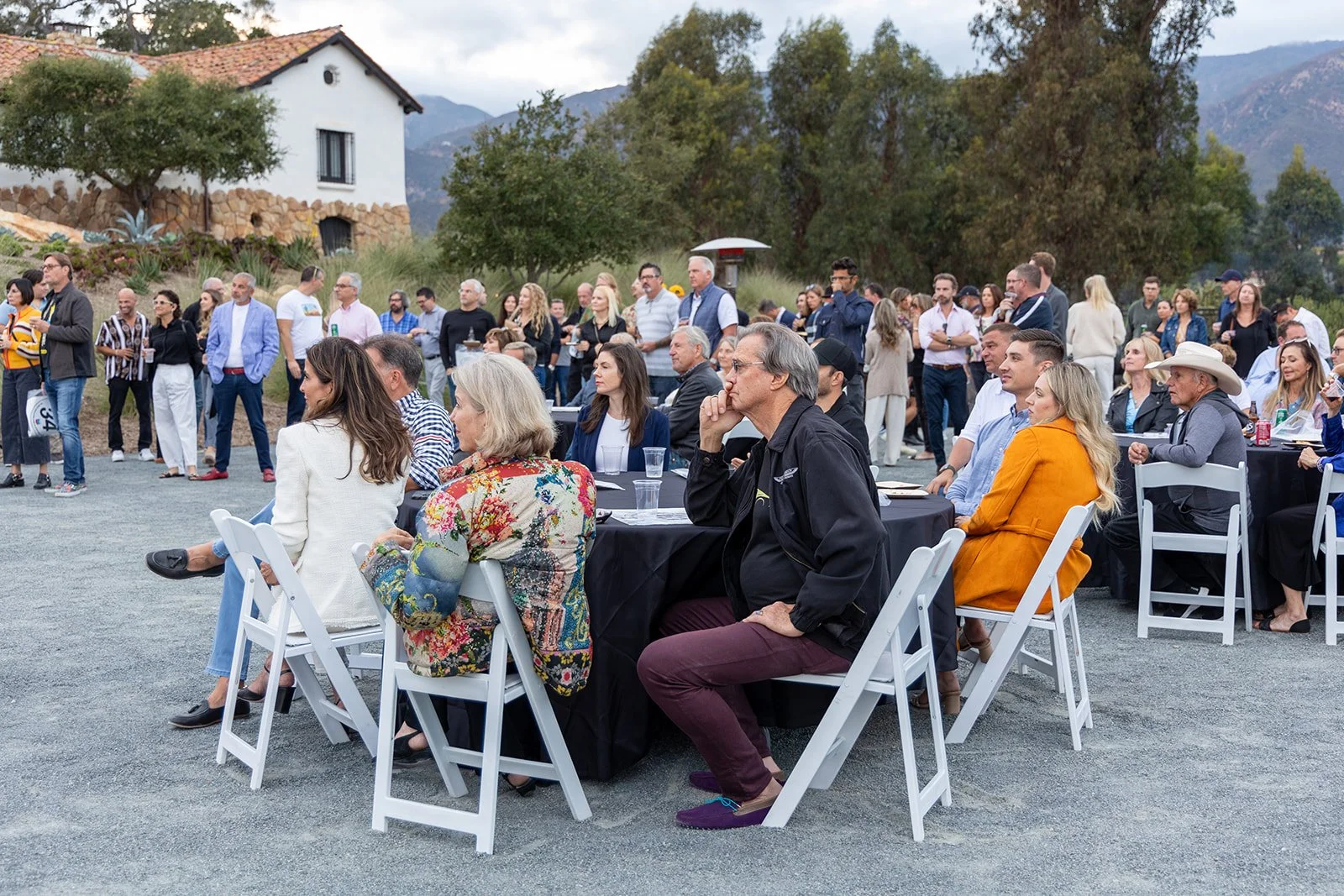 A group of people gathered outdoors, some seated at round tables with black tablecloths, others standing behind. The setting appears to be a casual outdoor event in a scenic area with mountains, trees, and a house in the background.