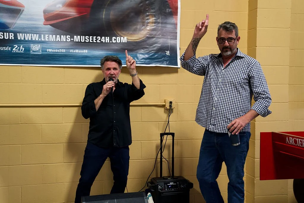 Two men standing in front of a yellow wall with a colorful banner behind them, one holding a microphone and the other raising his index finger.