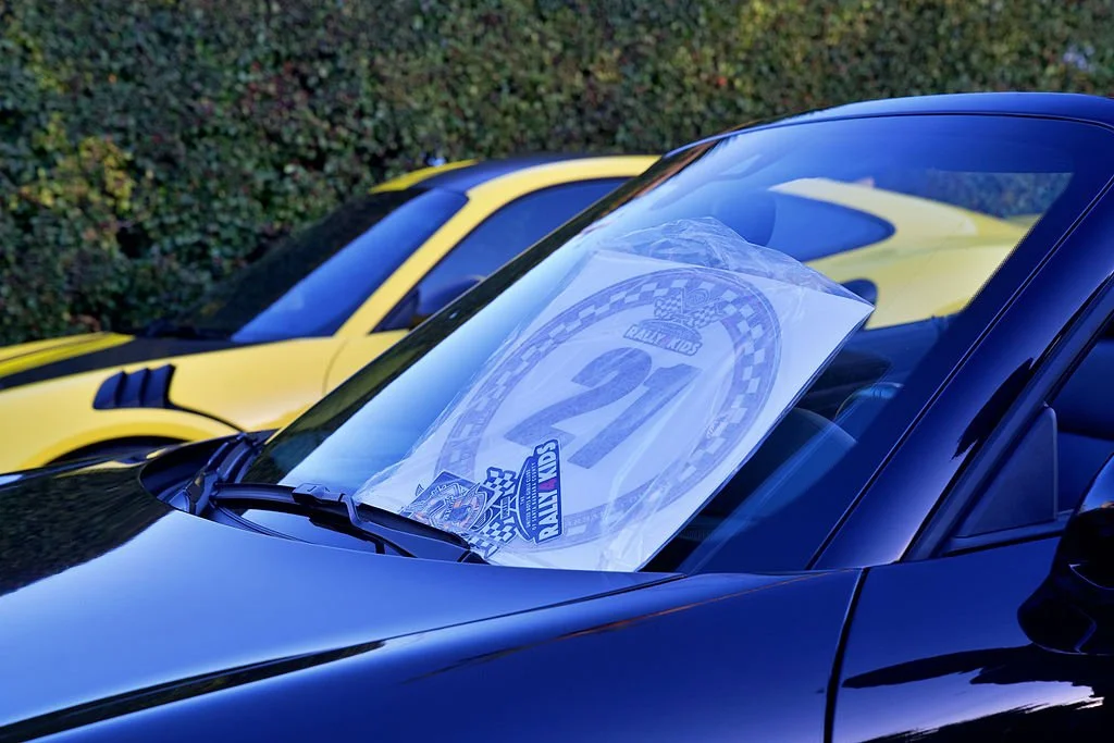Black sports car with a Race Kid poster on the windshield, and a yellow sports car in the background, parked outdoors.