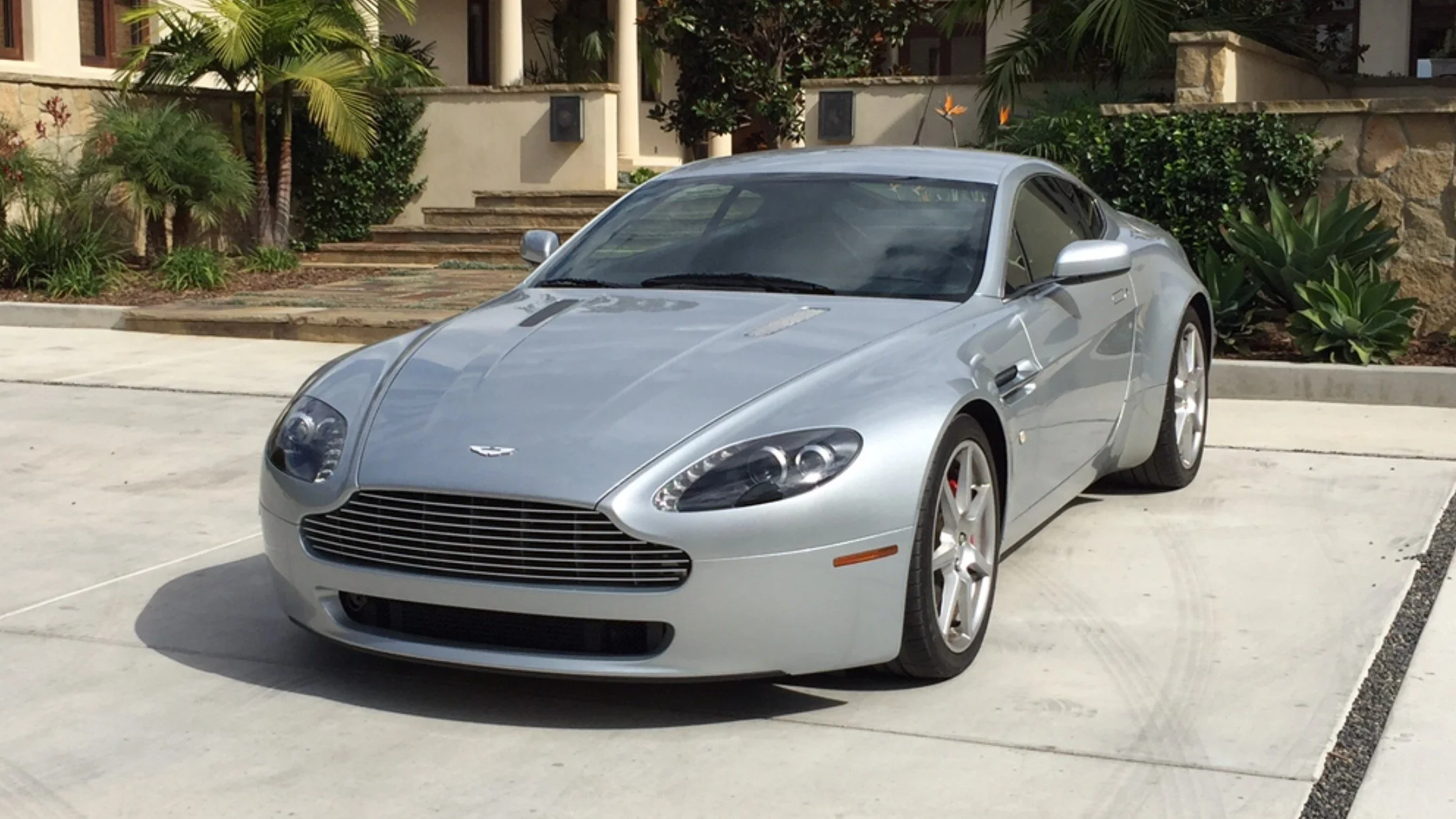 A silver Aston Martin sports car parked on a driveway in front of a house with landscaping that includes palm trees and succulent plants.