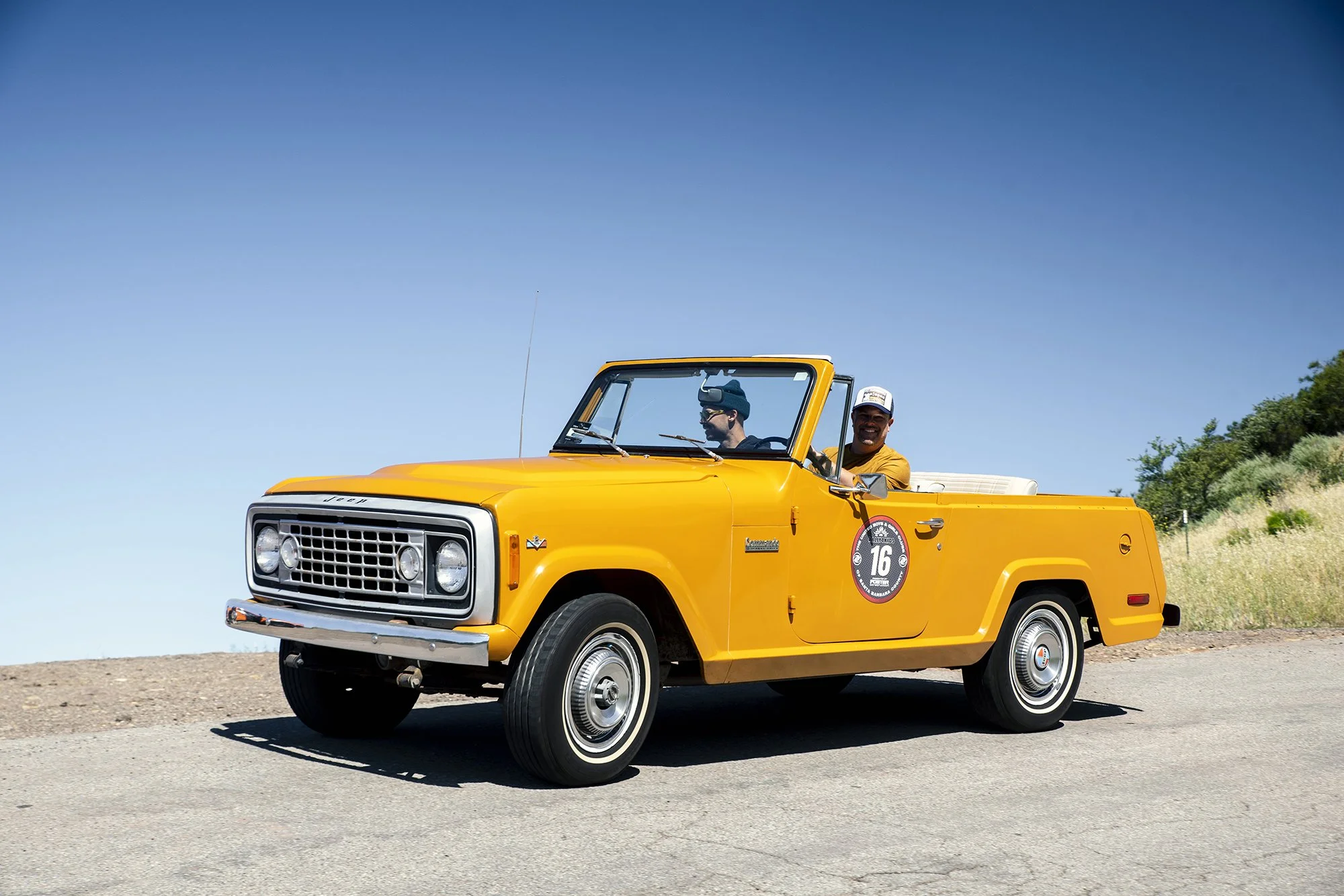 Yellow vintage Jeep vintage vehicle with two men inside, one wearing sunglasses and a cap, the other smiling, on a hill under a clear blue sky.