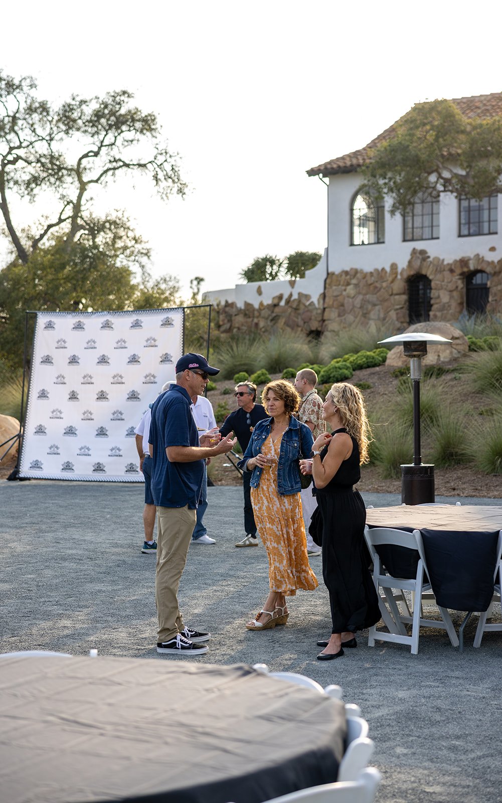 People socializing outdoors at a gathering during the daytime, with a backdrop of a white house with stone accents and a large window, surrounded by landscaping.