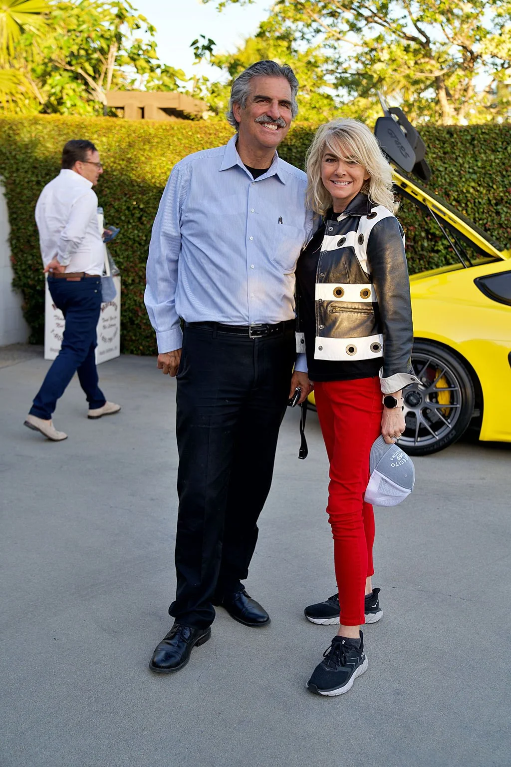 A man and woman standing together outdoors, smiling. The man wears a light blue button-up shirt and black pants, the woman wears a black and white leather jacket, red pants, and black sneakers. A yellow sports car with open gullwing doors and a racin