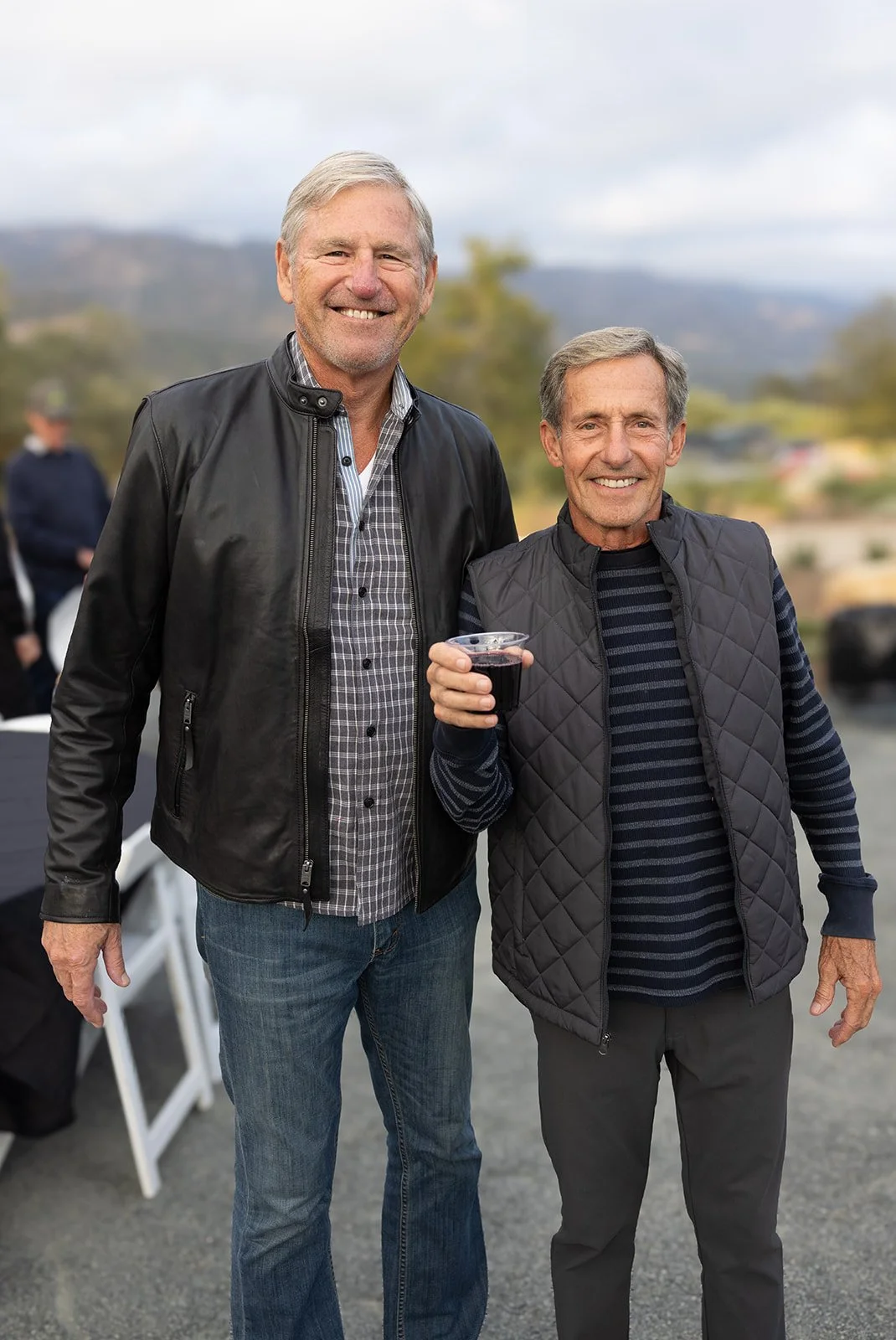 Two smiling older men standing outdoors, one holding a glass of red wine, with a blurred natural background.
