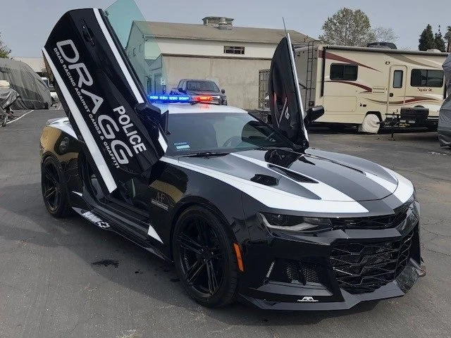 Black and white police sports car with front and rear doors open, displaying police decals and flashing blue and red siren lights.