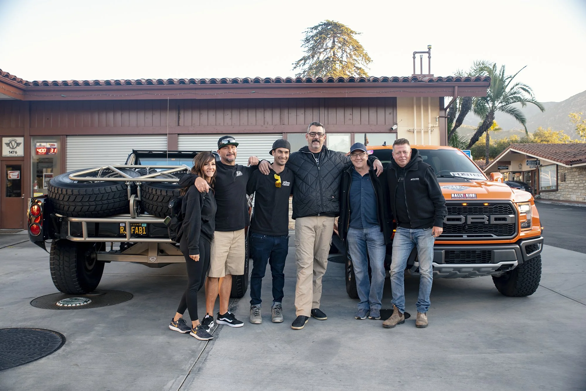 Group of six people standing together outdoors in front of a bright orange Ford pickup truck, with one person in a wheelchair, all smiling and embracing each other.