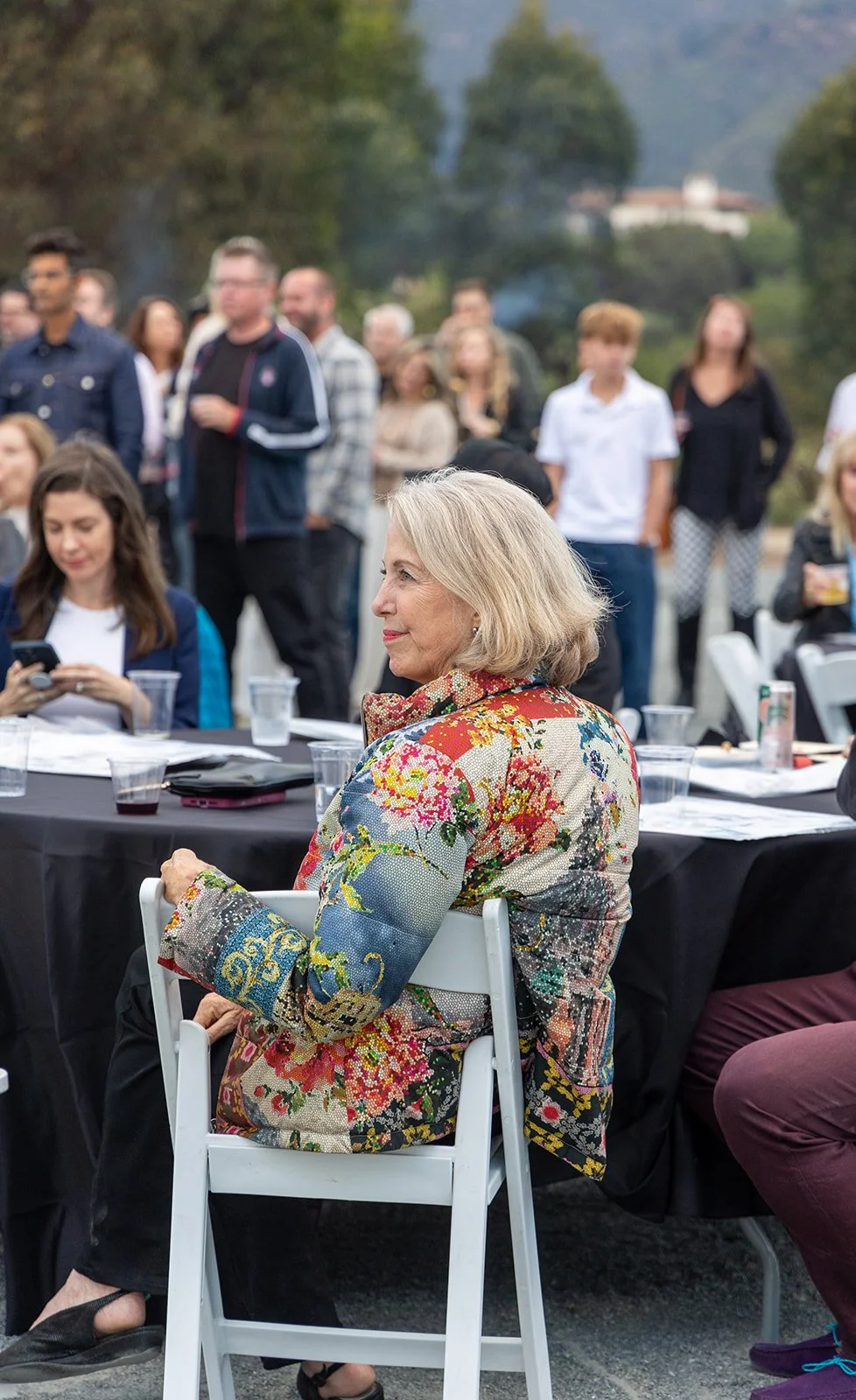 A woman with blonde hair wearing a colorful floral jacket sitting on a white chair at an outdoor event with a large crowd in the background.