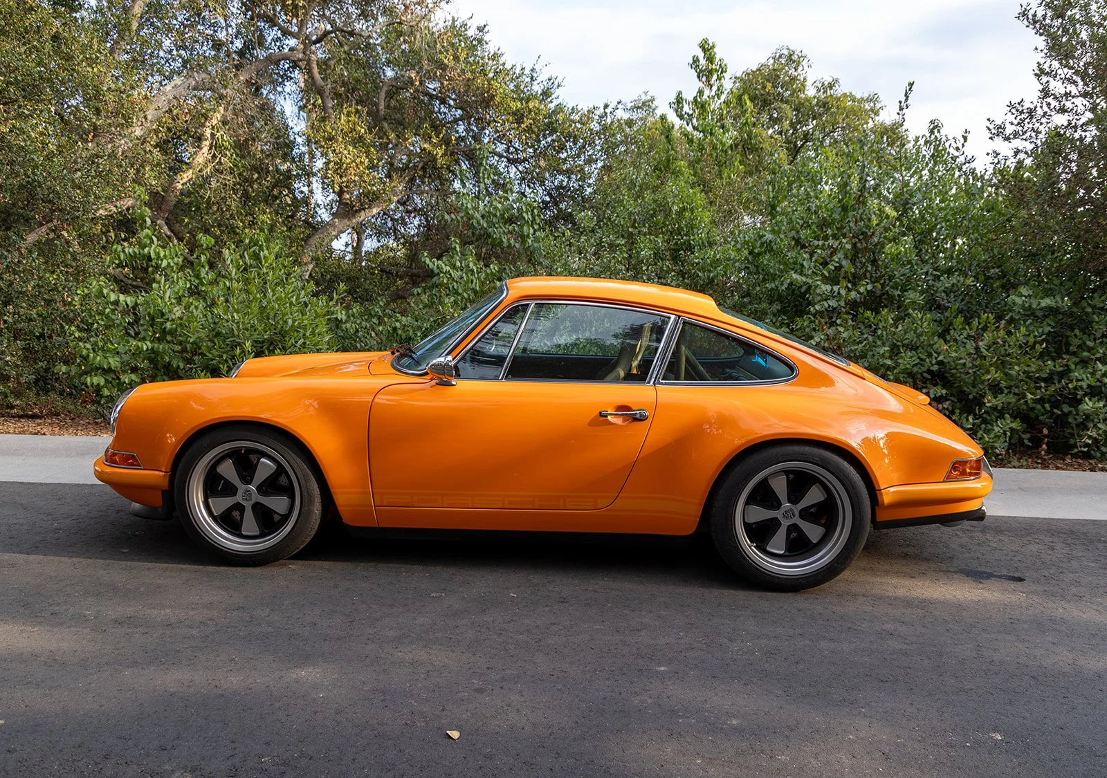 An orange classic Porsche sports car parked on a paved road with green bushes and trees in the background.